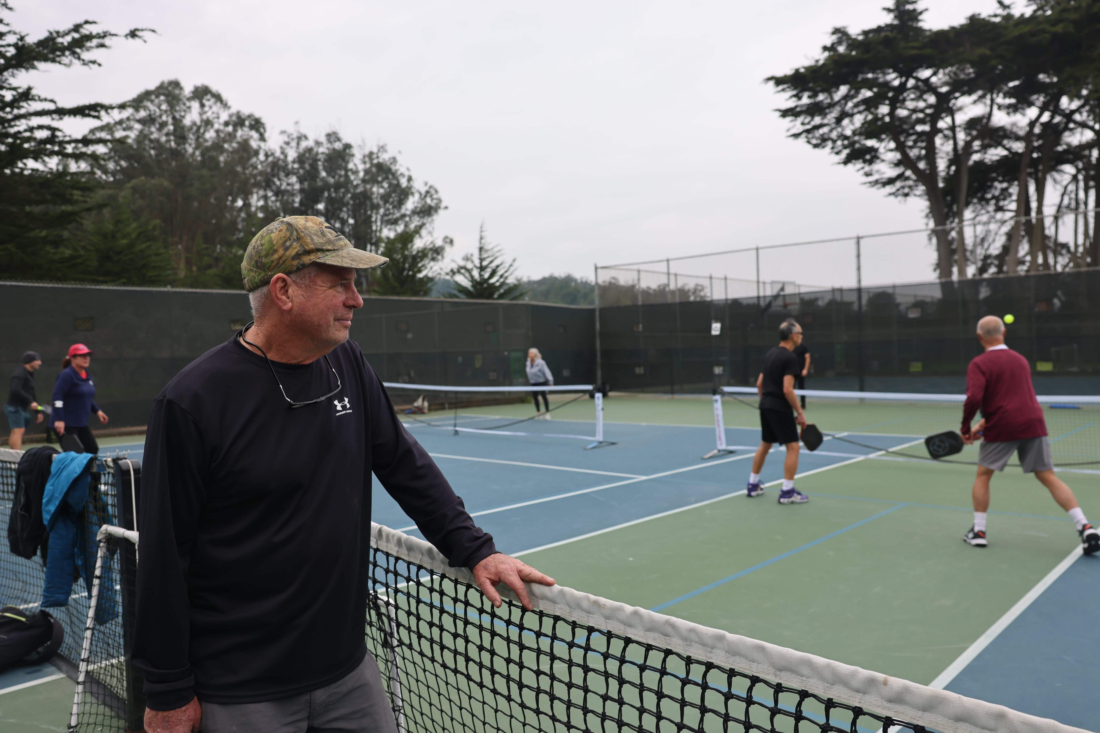 A man leans on a pickleball net, watching others play pickleball on a cloudy day.
