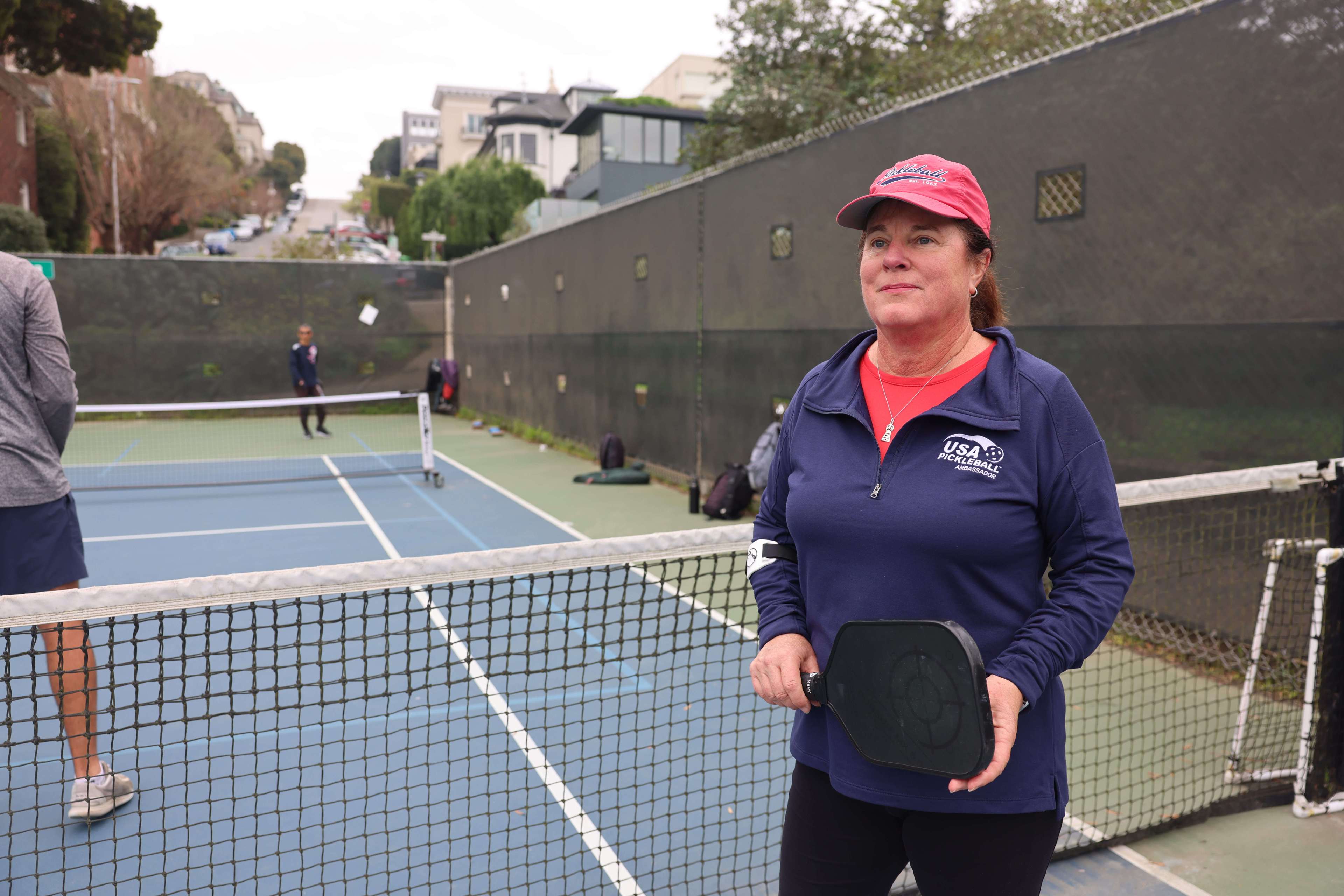 A woman stands by a pickleball net with a paddle, wearing a cap and sportswear, with players and courts in the background.
