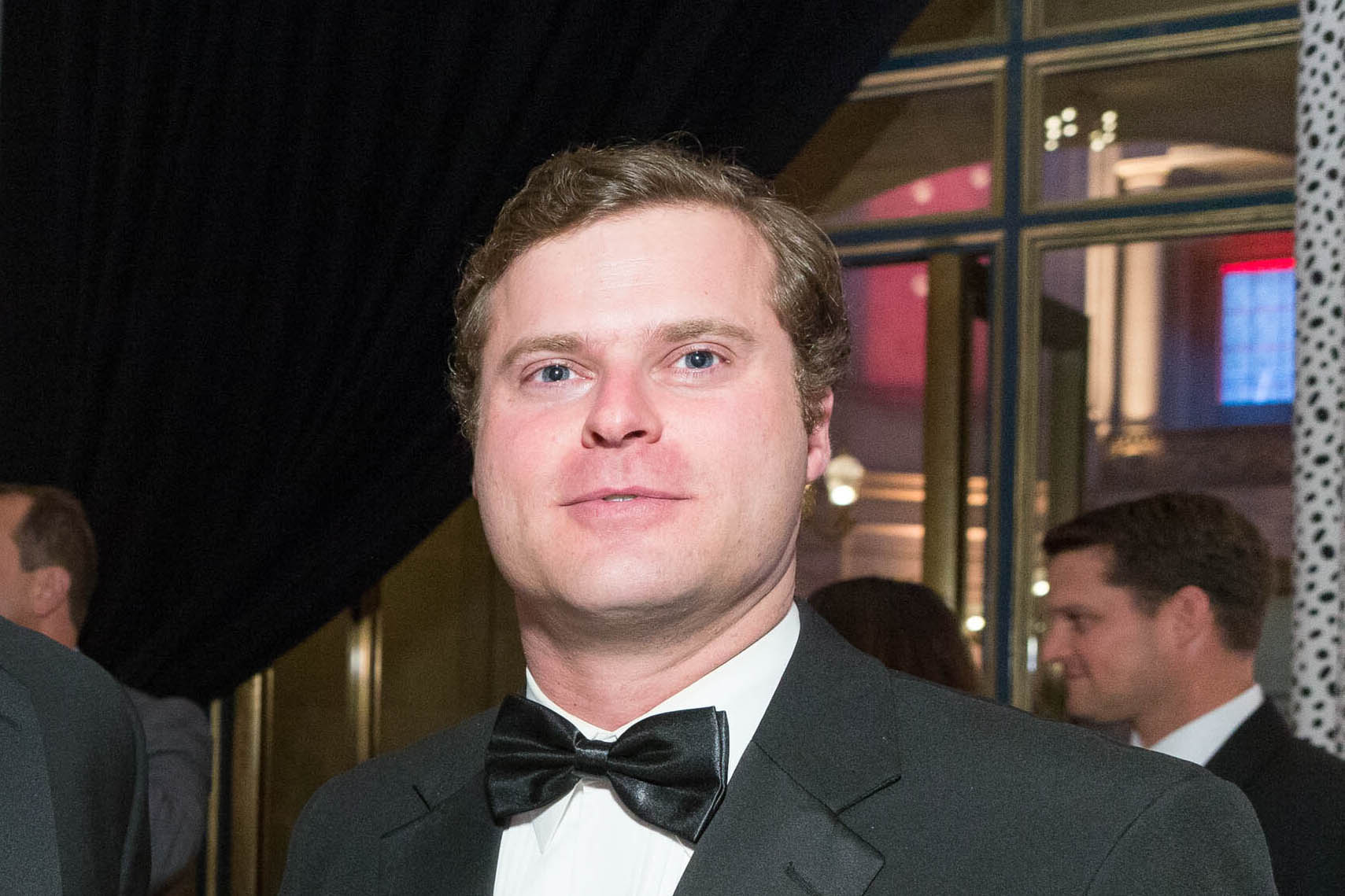 A man in formal attire with a black bow tie stands indoors, surrounded by people. The background includes elegant lighting and architectural details.