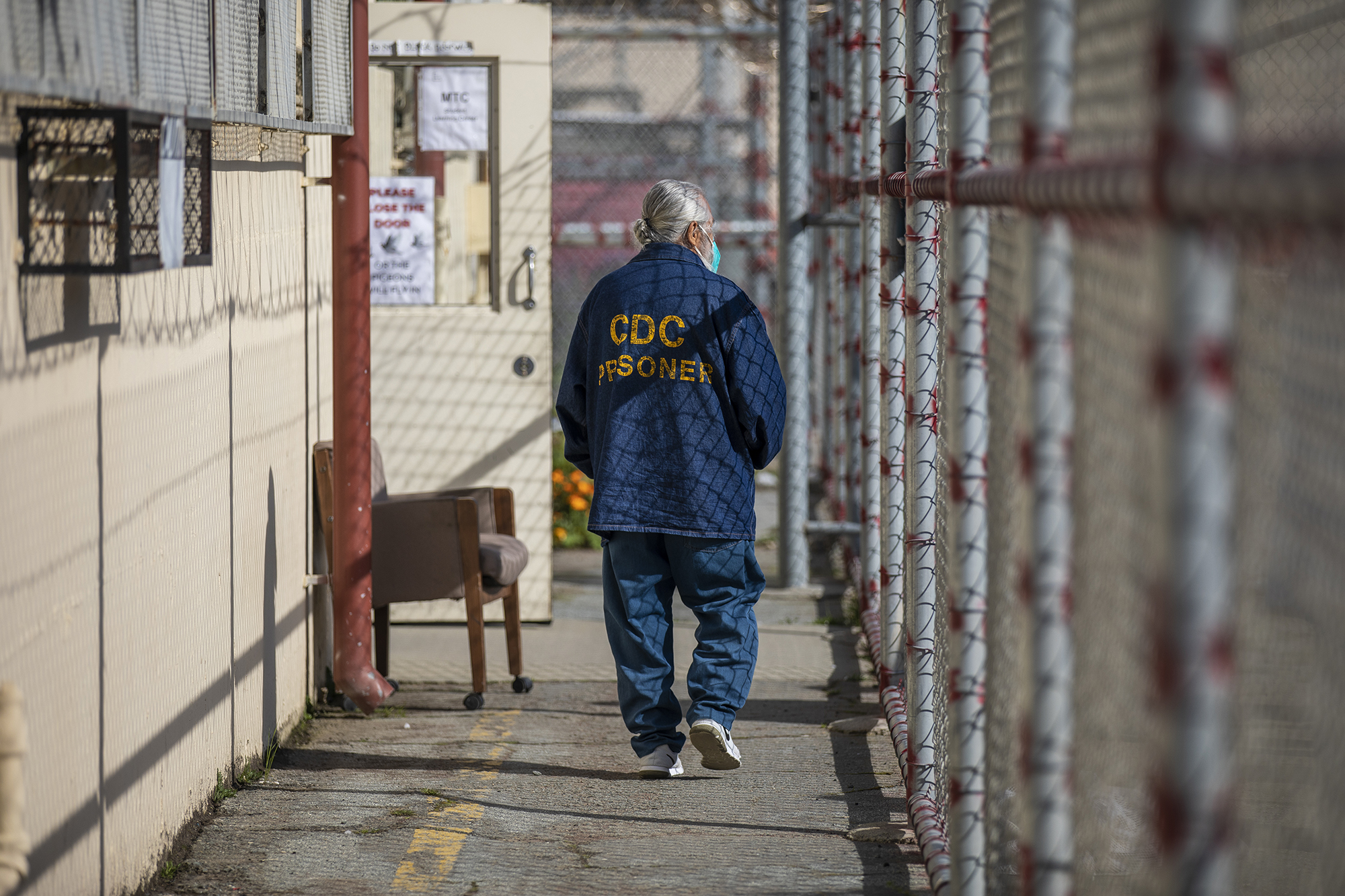 Man with gray pony tail wearing blue pants and a blue shirt with the words CDC Prisoner on it walks next to a chain link fence