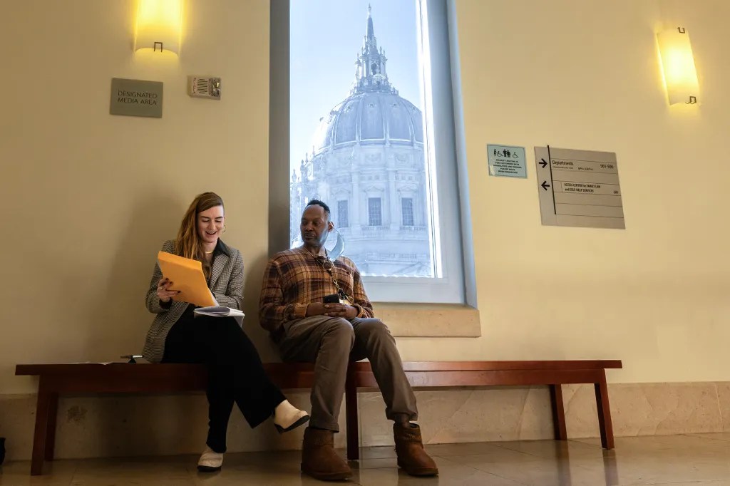 Man and woman sit on bench in hallway in front of window showing government building rotunda