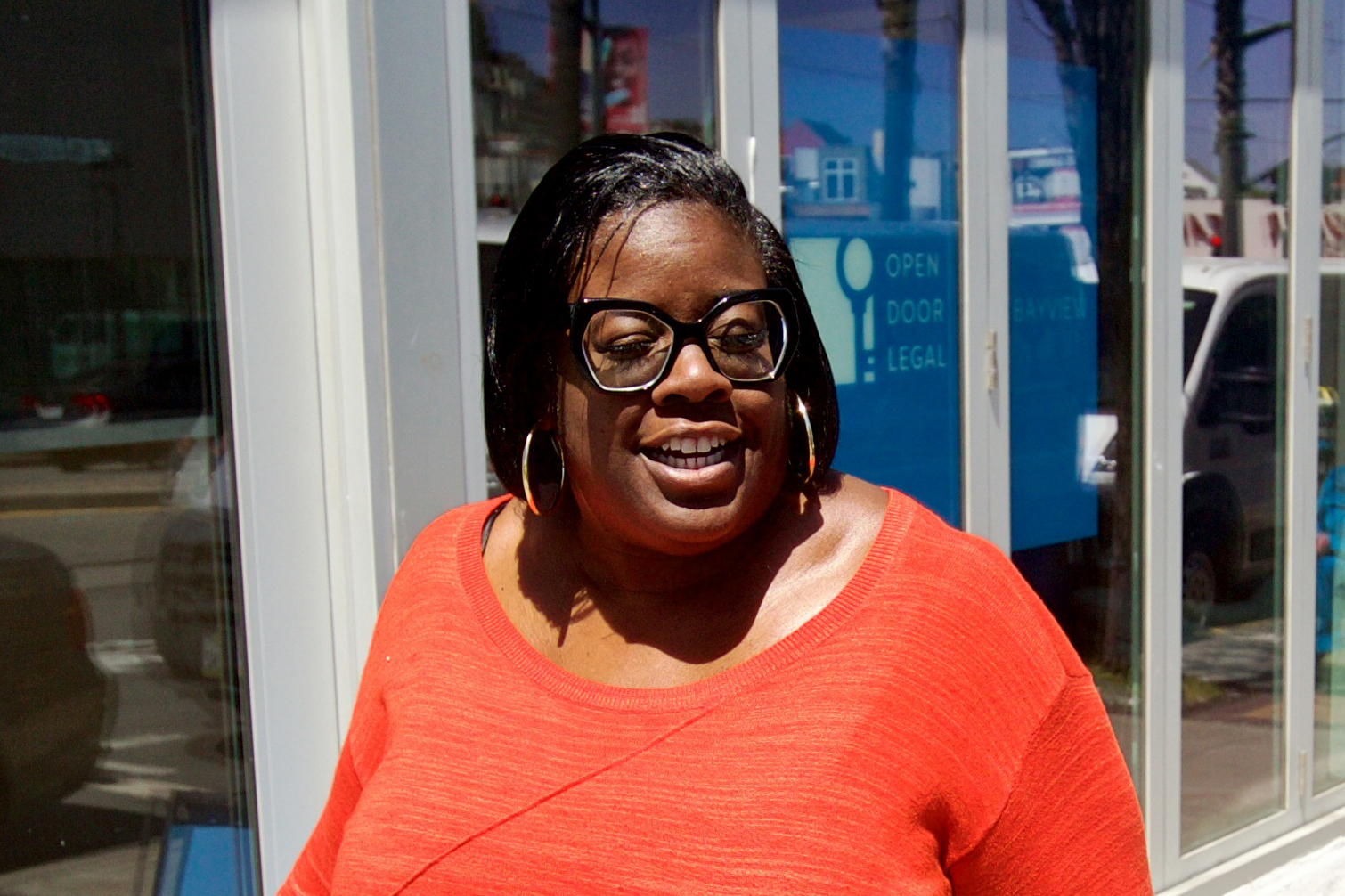 A smiling woman in an orange top with glasses and hoop earrings stands in front of a window with blue signage.