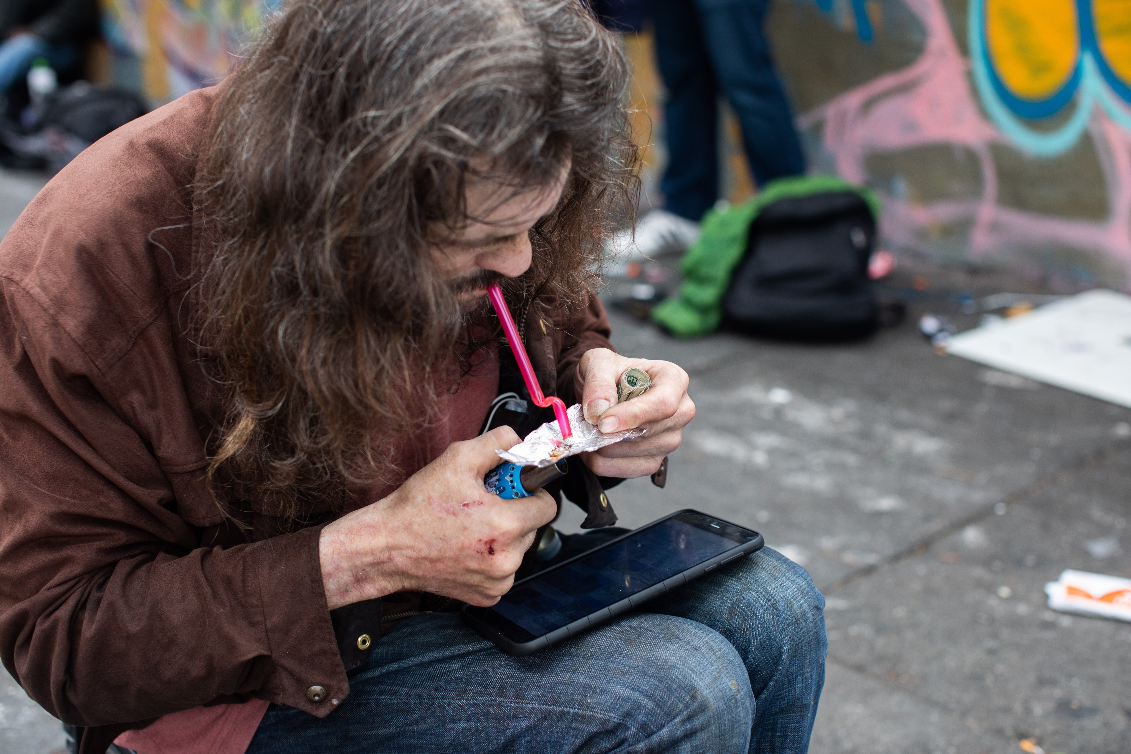 A man is hunched over, using a straw on a device in his hand, with graffiti and items in the background.