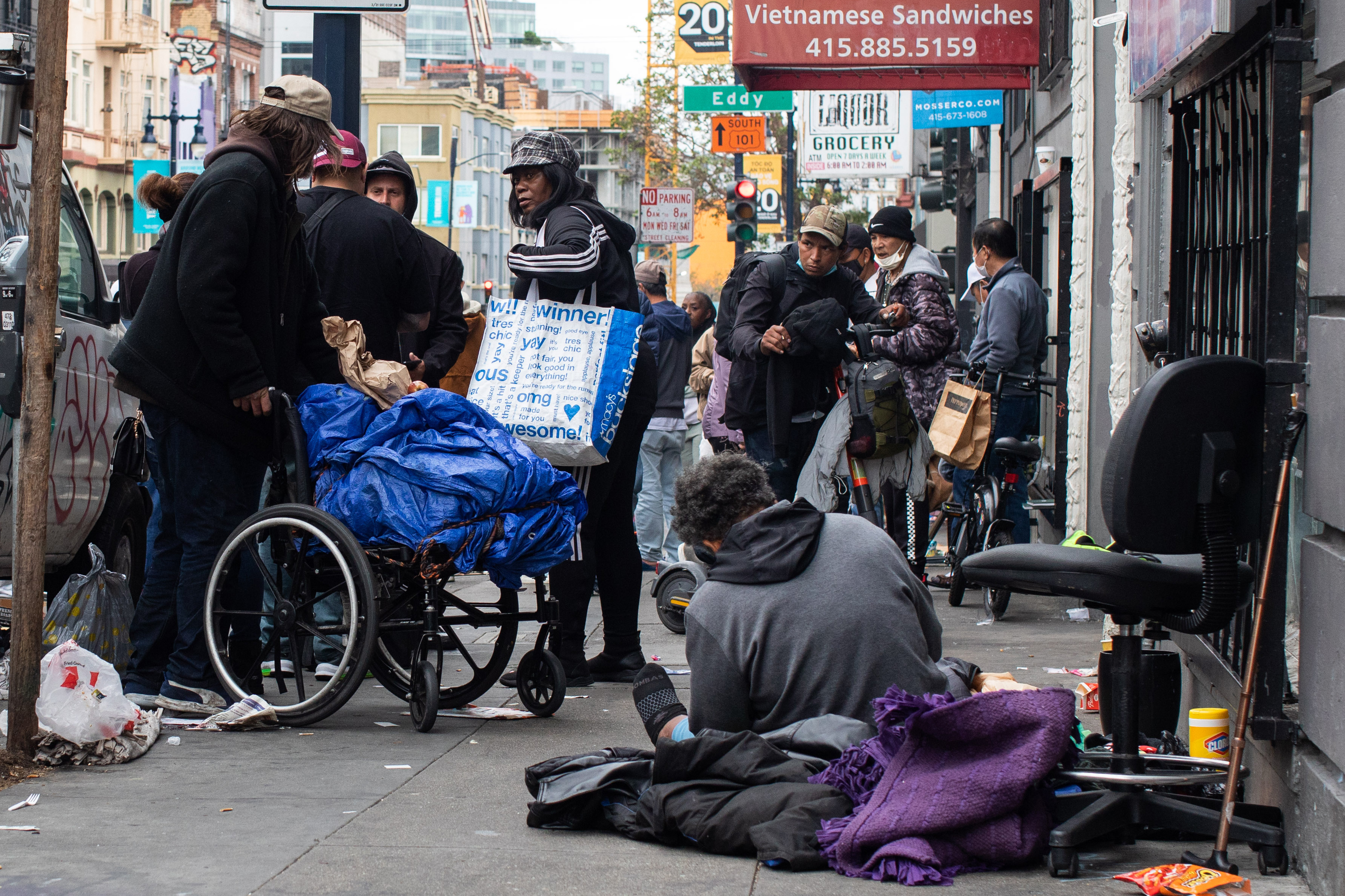 An urban street scene with people gathered, some standing, one seated on the ground, amidst litter and a wheelchair with belongings.
