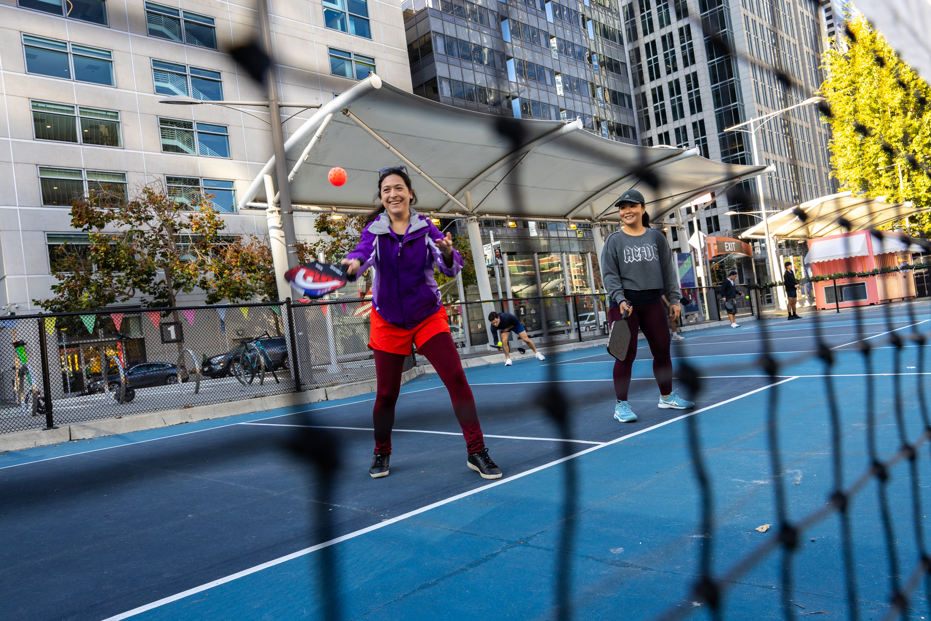 Two people playing pickleball on an urban court, smiling, with buildings in the background.