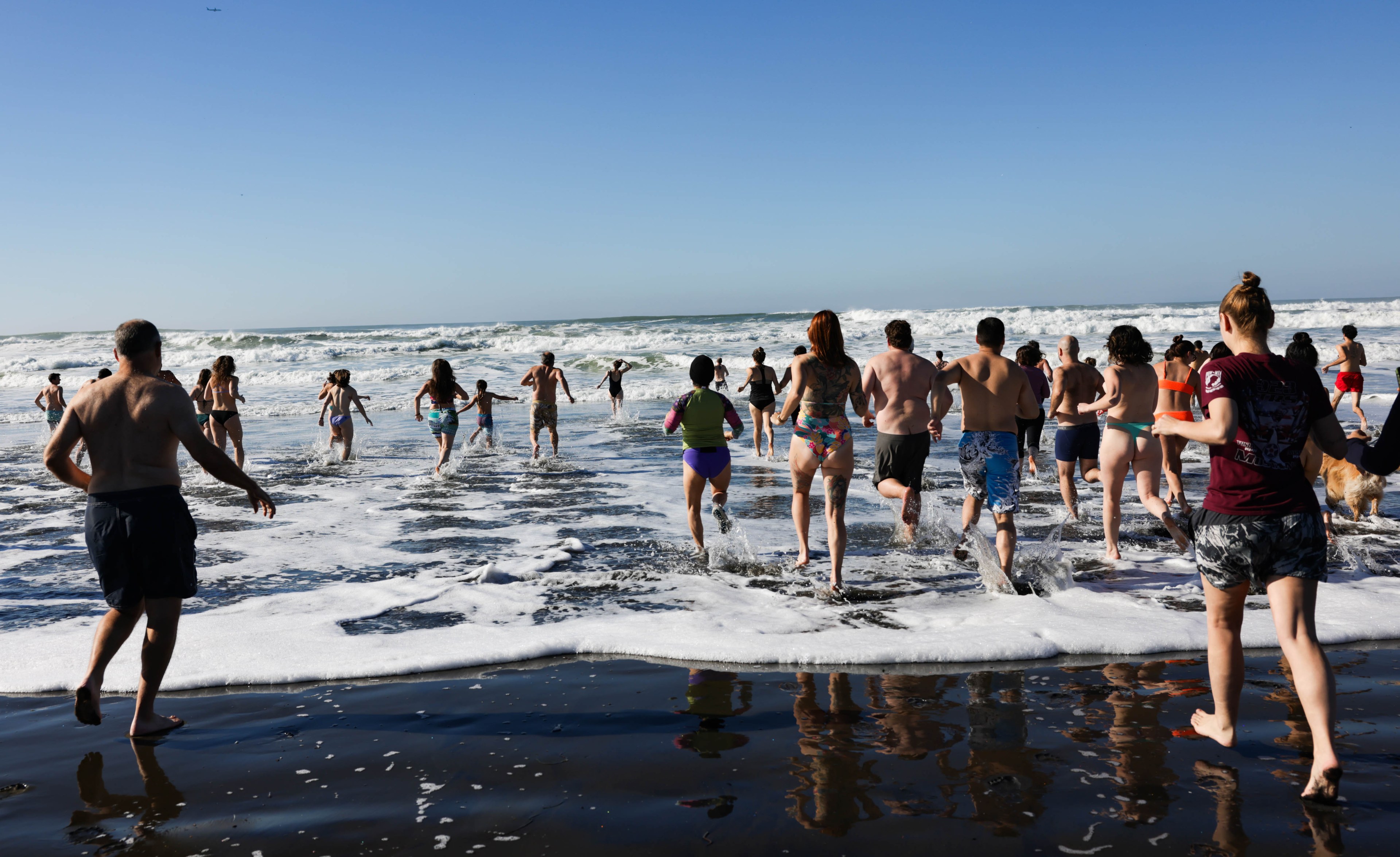 A group of people in swimsuits run into the ocean, splashing through shallow waves on a sunny day, with a clear blue sky above.