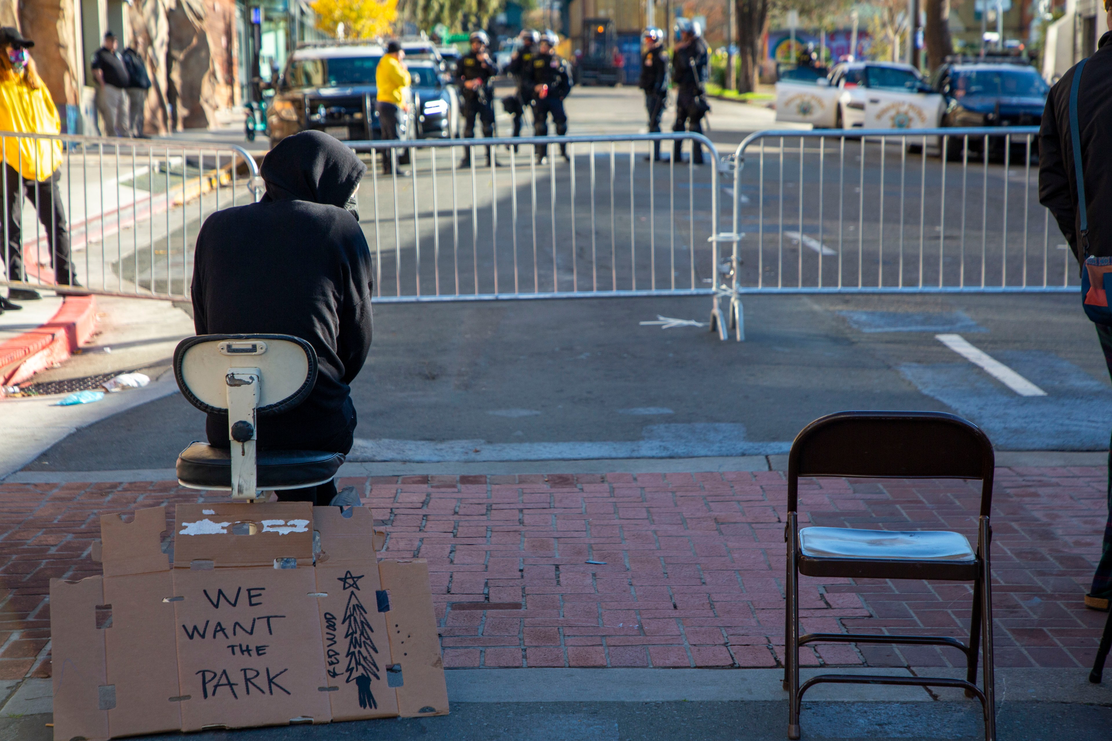 A person wearing a dark hooded sweatshirt sits in a chair in front of police on the other side of a metal barricade; behind the person, a cardboard sign reads &quot;we want the park.&quot;