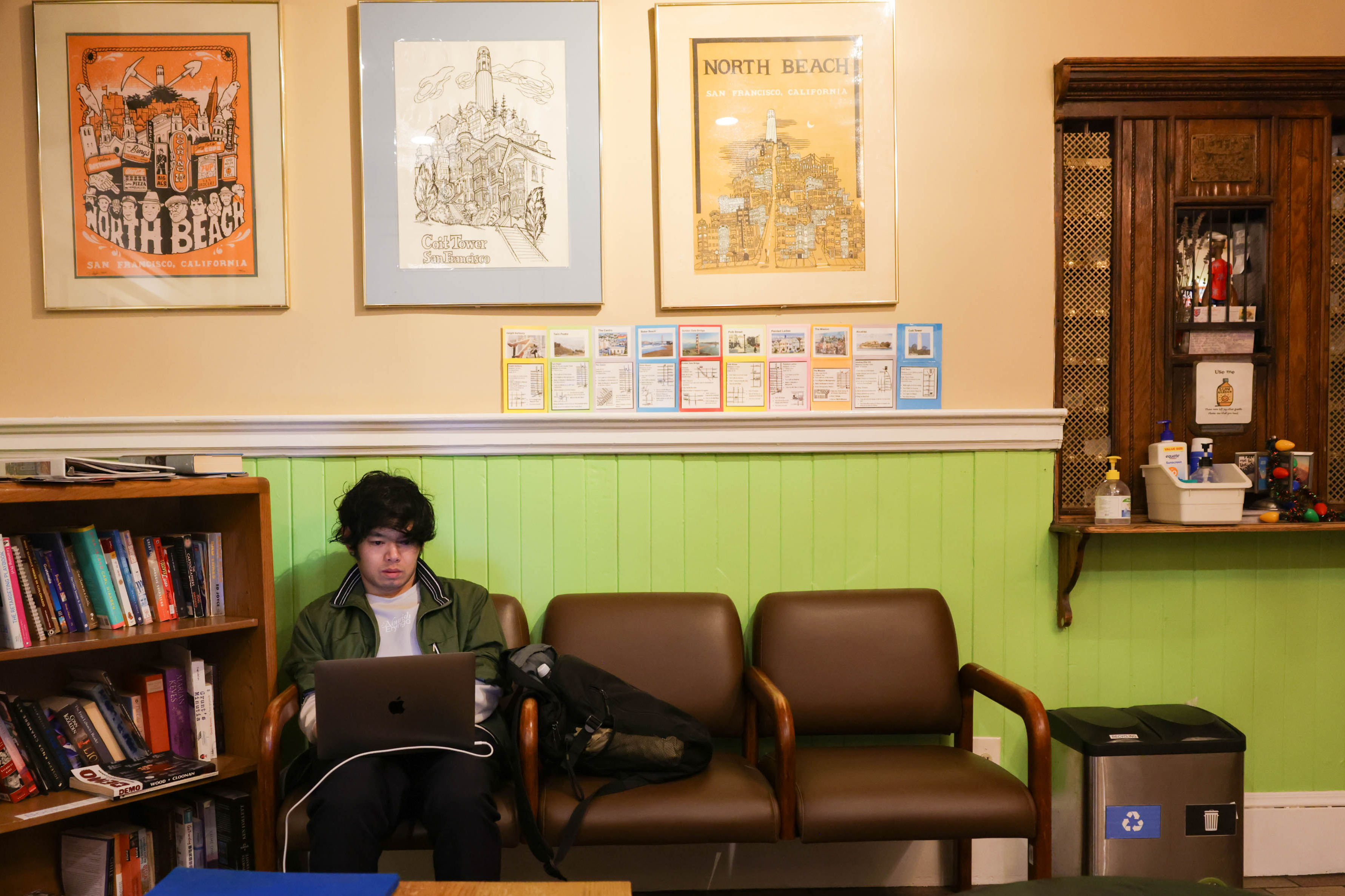 A person is seated between two chairs, using a laptop near a bookshelf, under posters, in a room with green wainscoting.