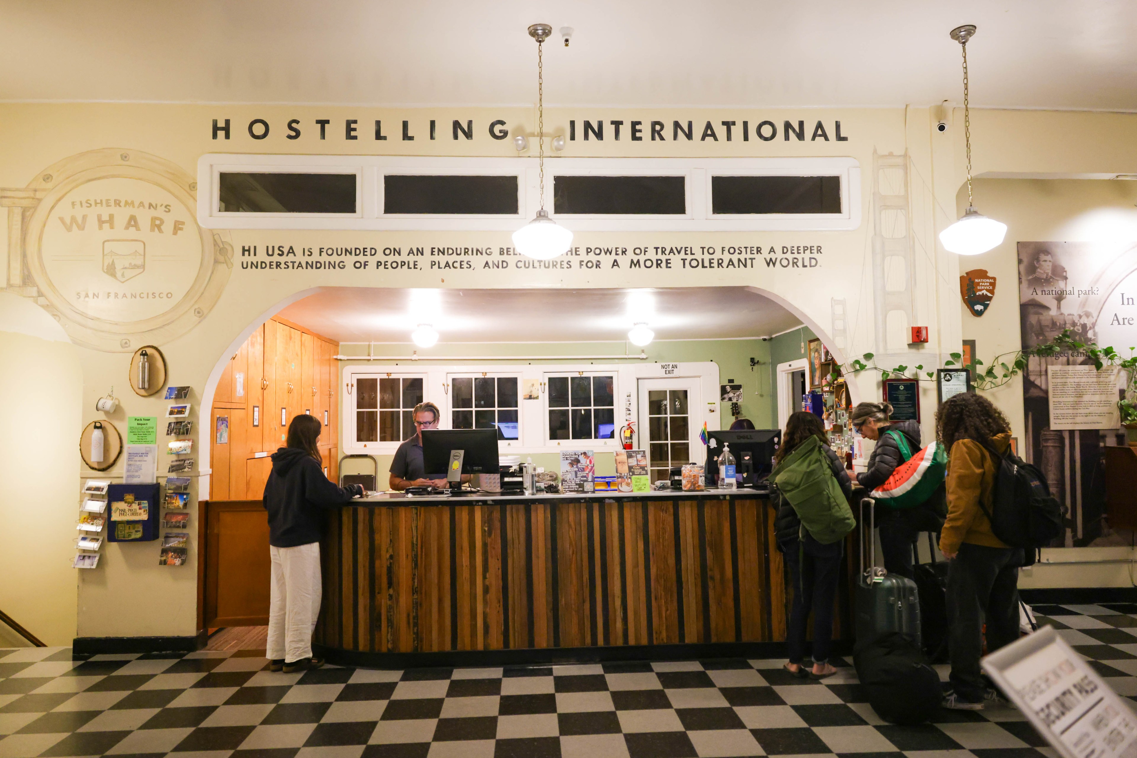 Hostel reception with guests, sign &quot;HOSTELLING INTERNATIONAL,&quot; and a mission statement for tolerance.