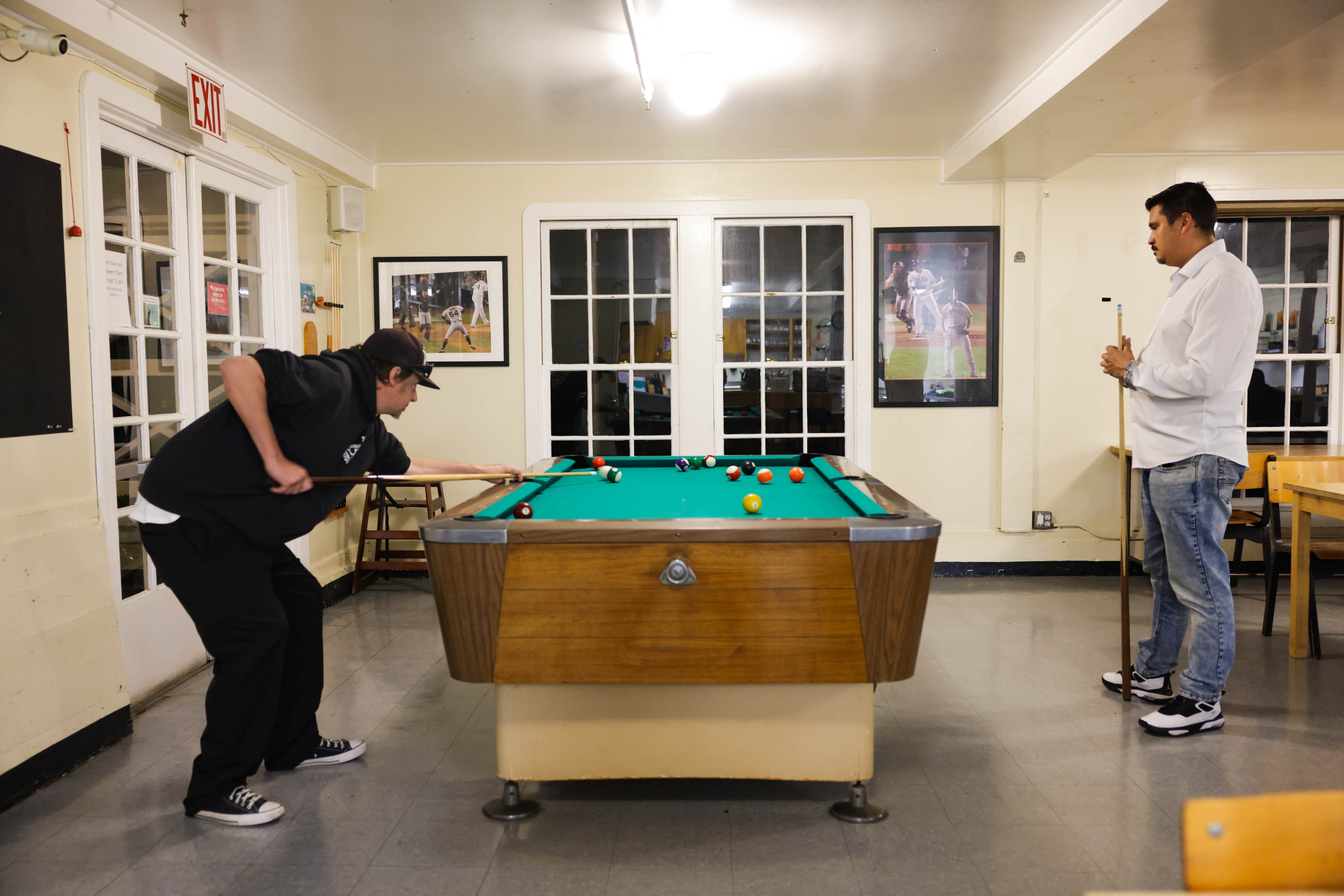 Two men are playing pool in a room with sports photos on the walls.