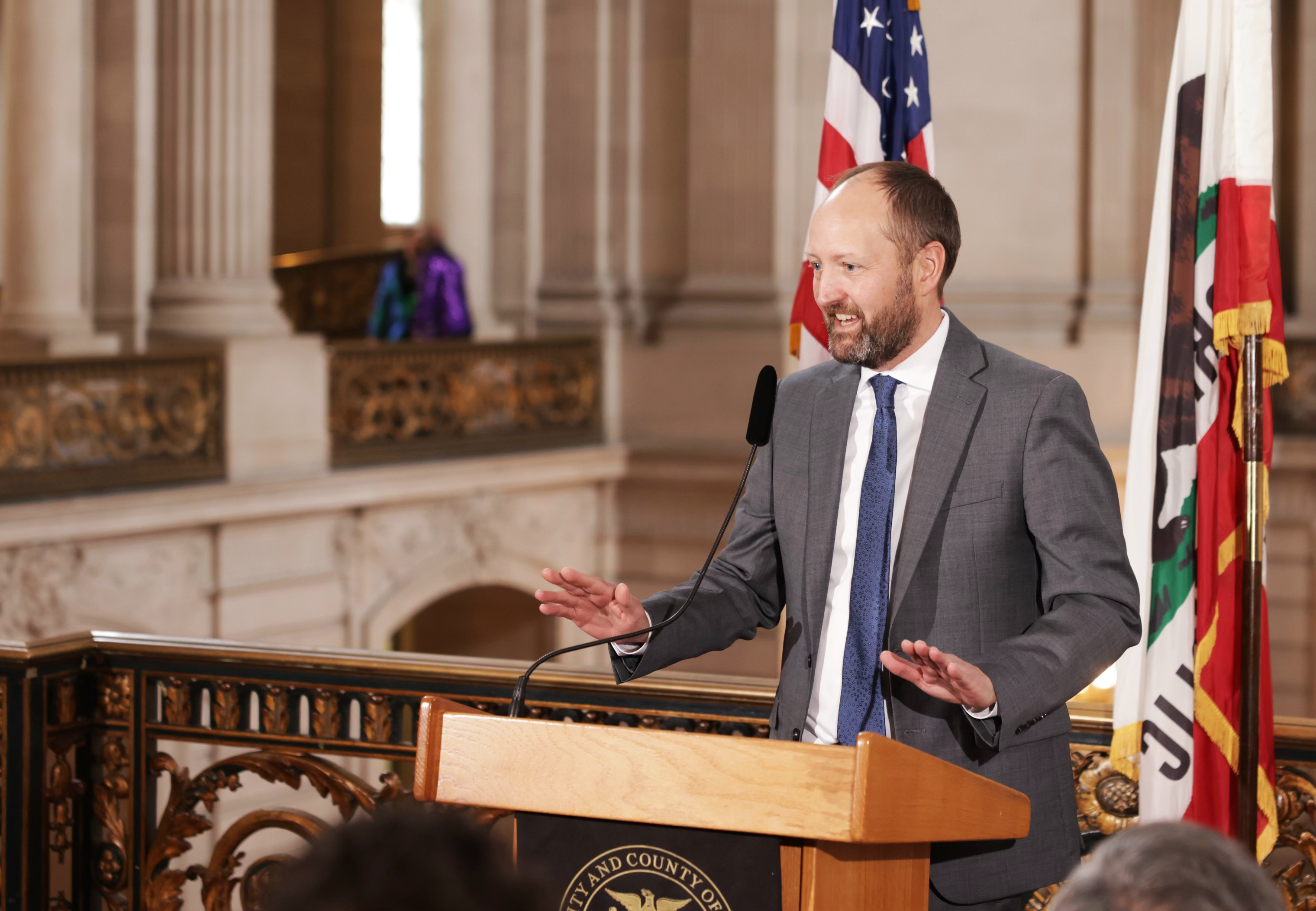 A man in a grey suit speaks behind a podium in front of an American and Californian flag at San Francisco City Hall. 