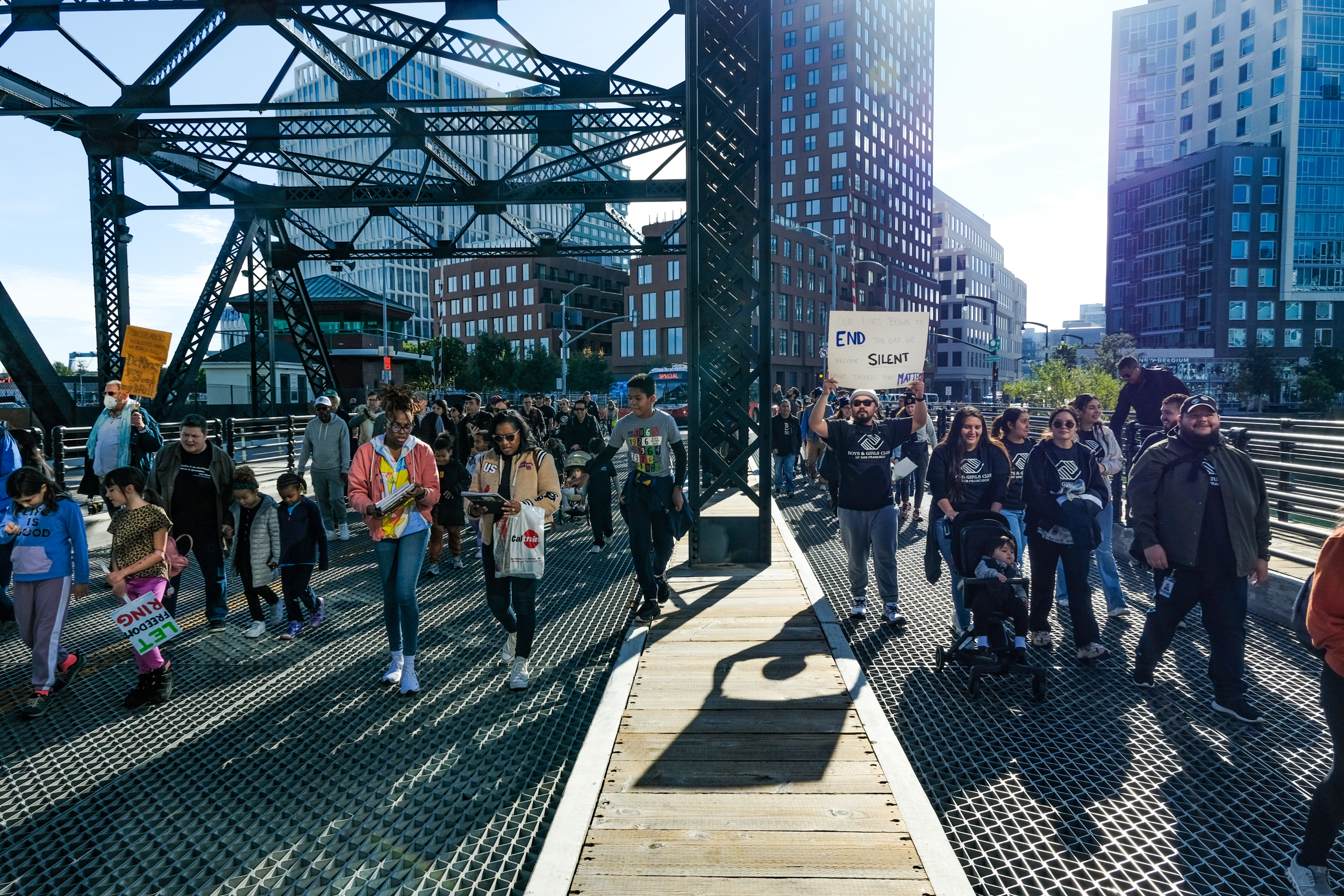 A diverse crowd walks across a metal bridge in a city, some holding signs, with urban buildings in the background.