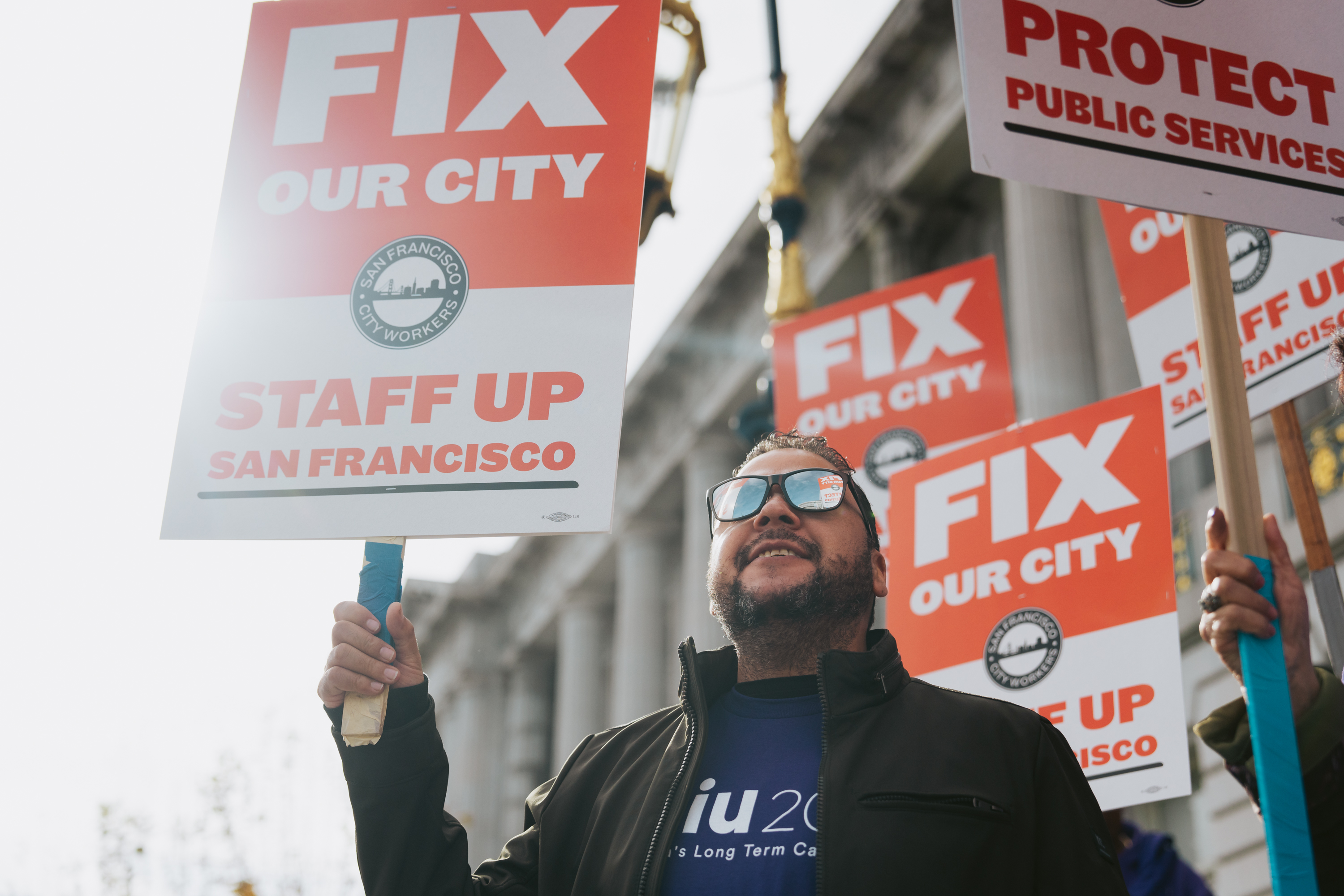 A person in sunglasses at a rally, holding signs that say &quot;FIX OUR CITY&quot; and &quot;STAFF UP SAN FRANCISCO.&quot;