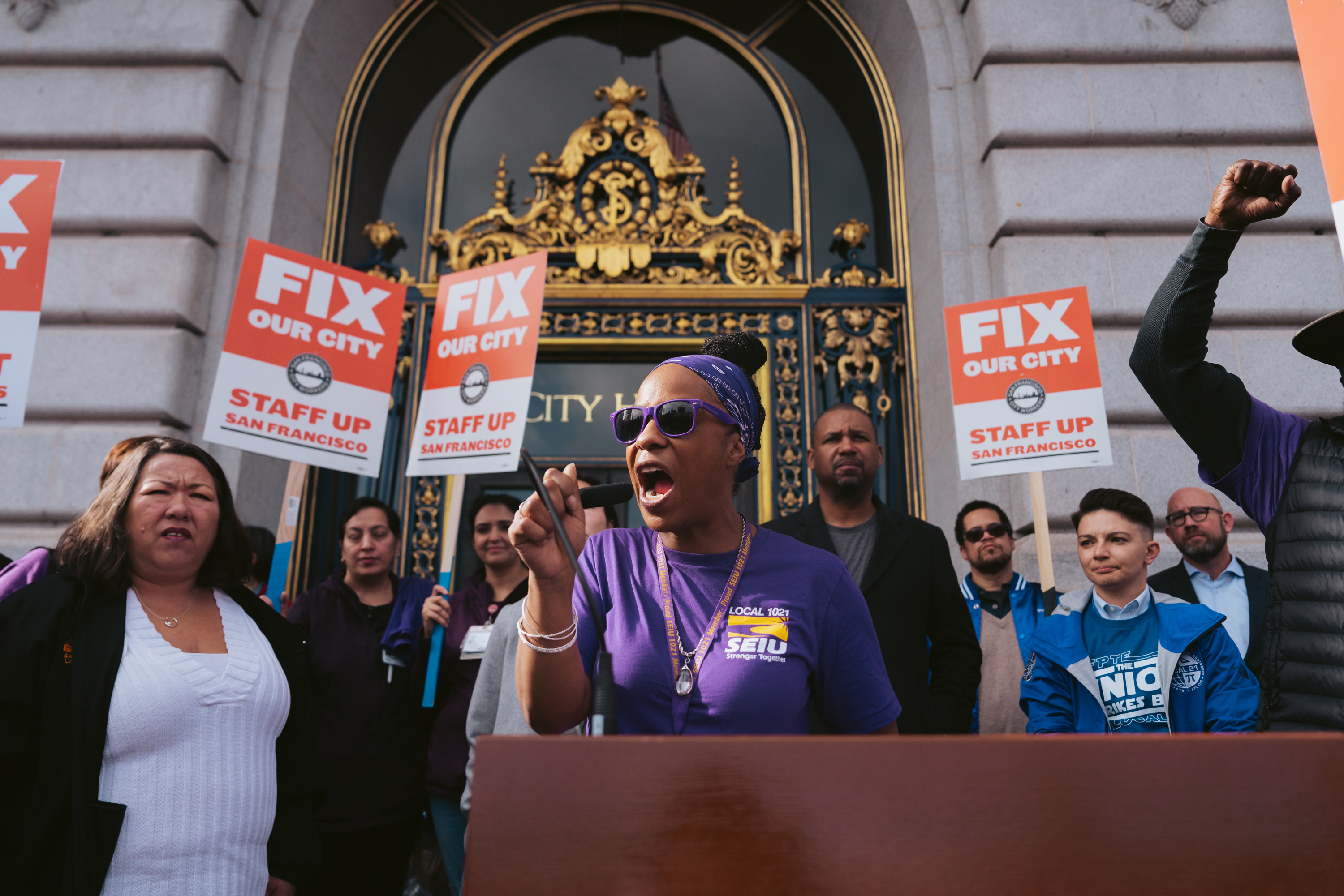 A group protests with signs reading &quot;FIX OUR CITY,&quot; a woman in purple speaks passionately into a microphone.