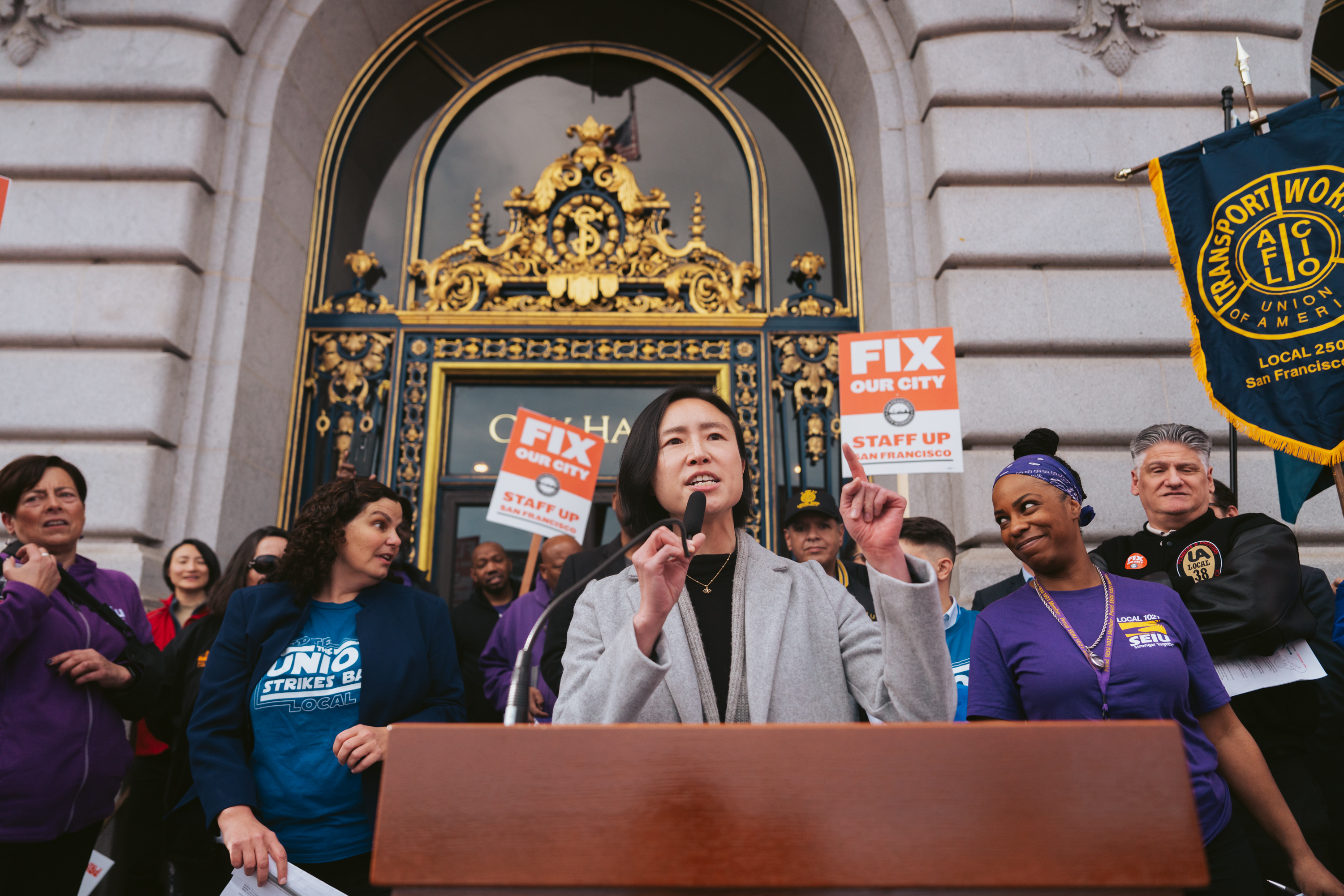 A person speaks at a podium before a group, some with union signs, outside an ornate building.