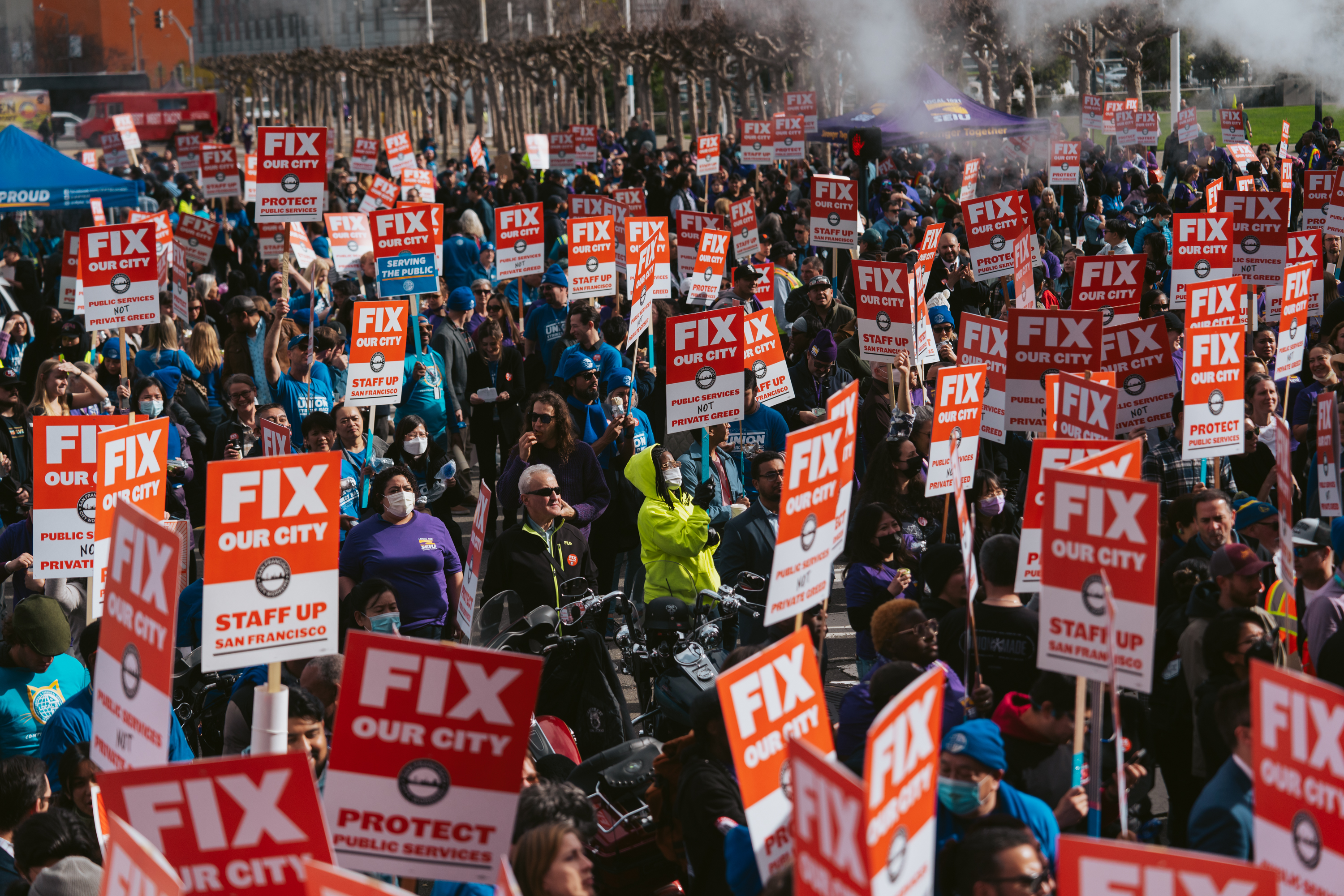 A large crowd holding signs that read &quot;FIX OUR CITY&quot; during a protest or rally, with many wearing blue and purple.
