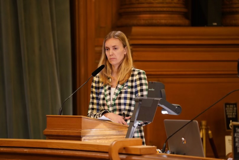 Mayor London Breed's budget director Anna Dunning, wearing a checked blazer, speaks at a podium in the Board of Supervisors chambers at City Hall.