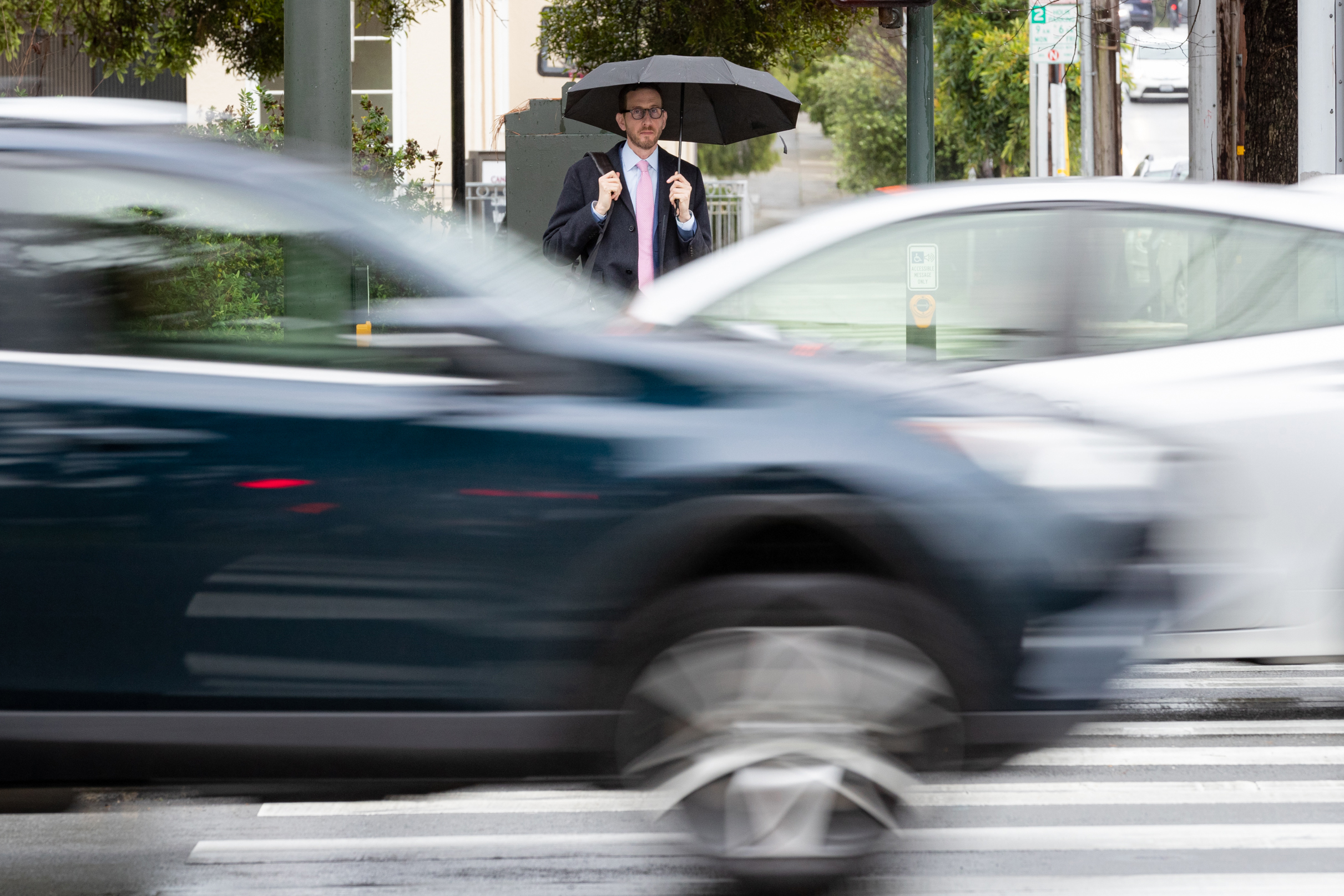 A man stands at a crosswalk as two cars drive past.
