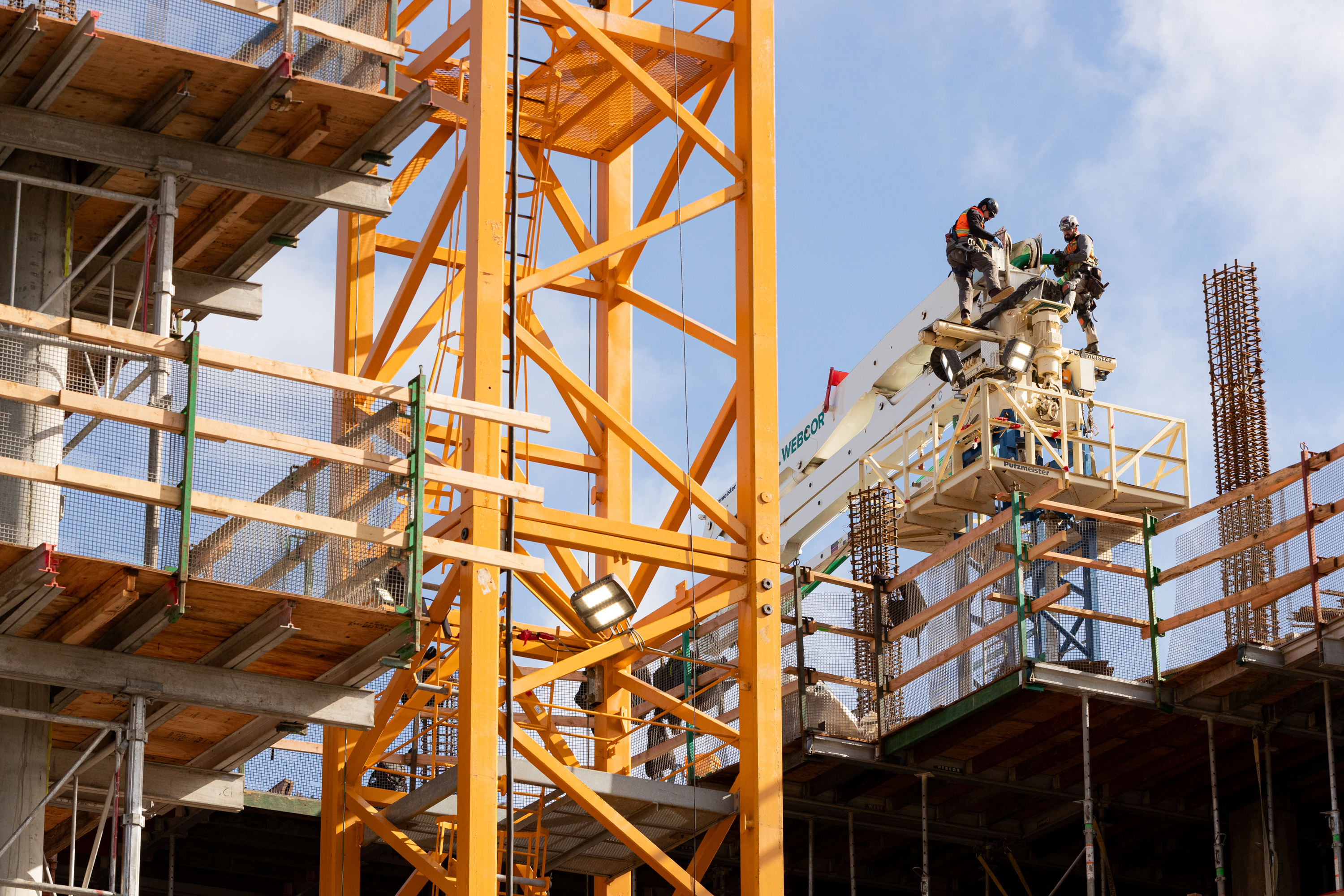 Two construction workers are on a lift at a building site. They're surrounded by orange steel beams and scaffolding under a blue sky.