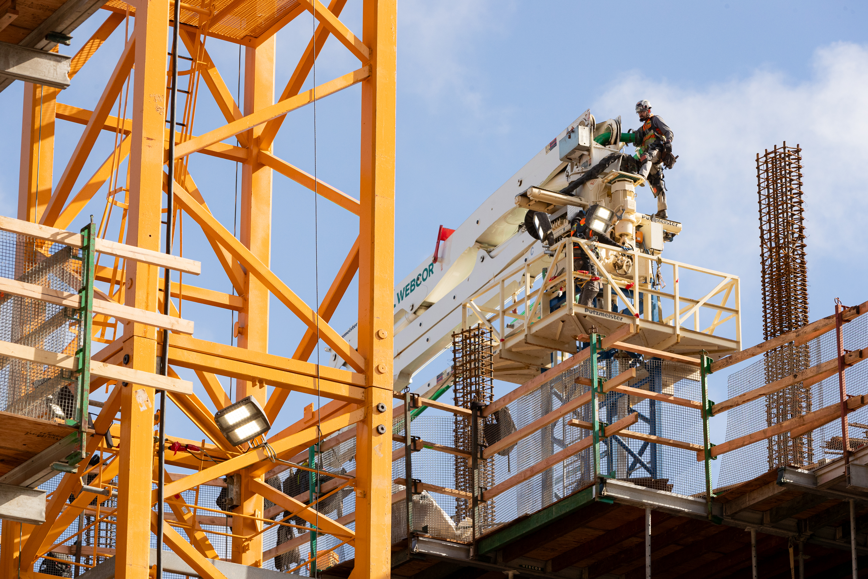 A construction site with a worker on a platform attached to a crane, surrounded by steel beams and scaffolding, under a clear blue sky.