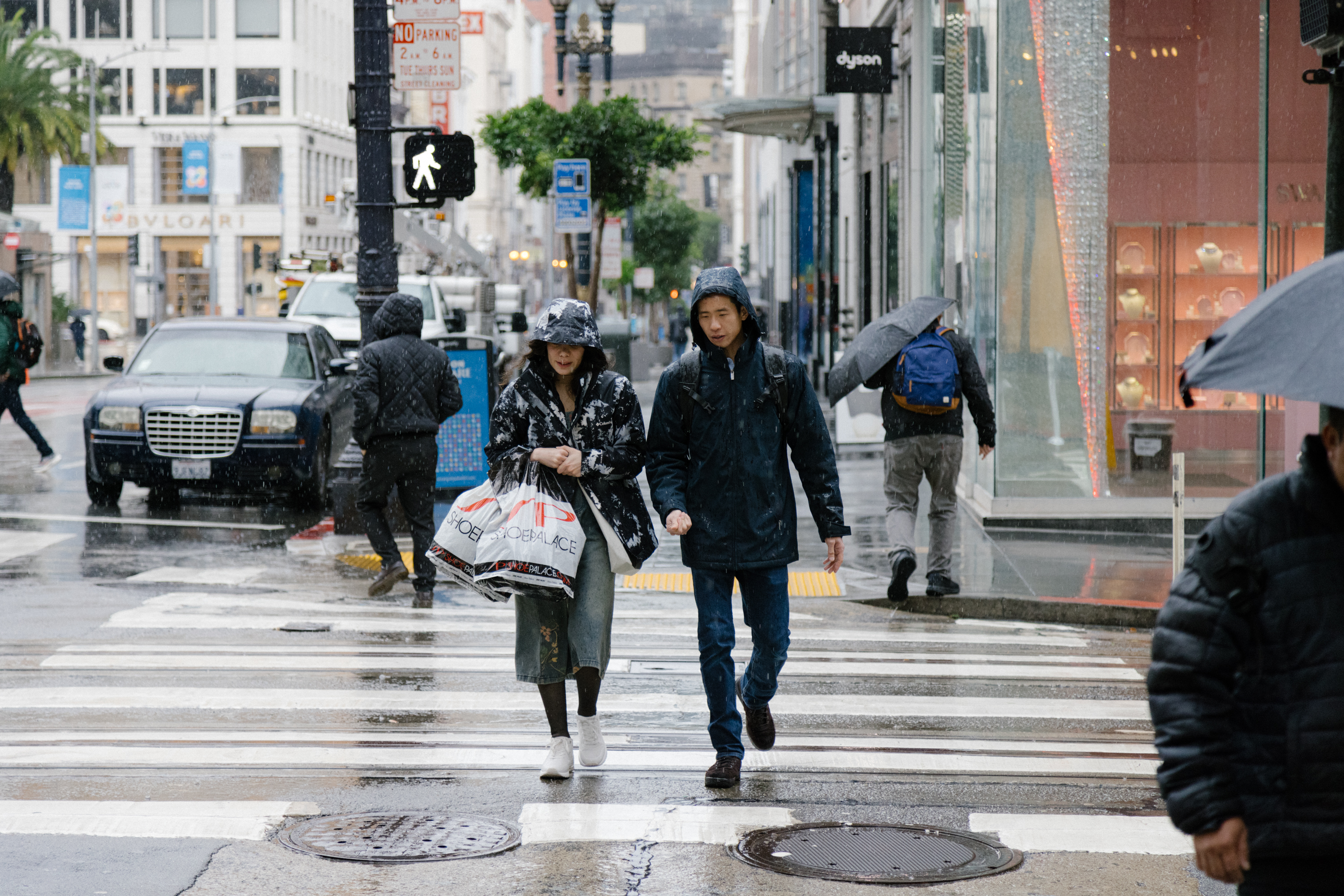 People in rain jackets and with umbrellas walk across wet street