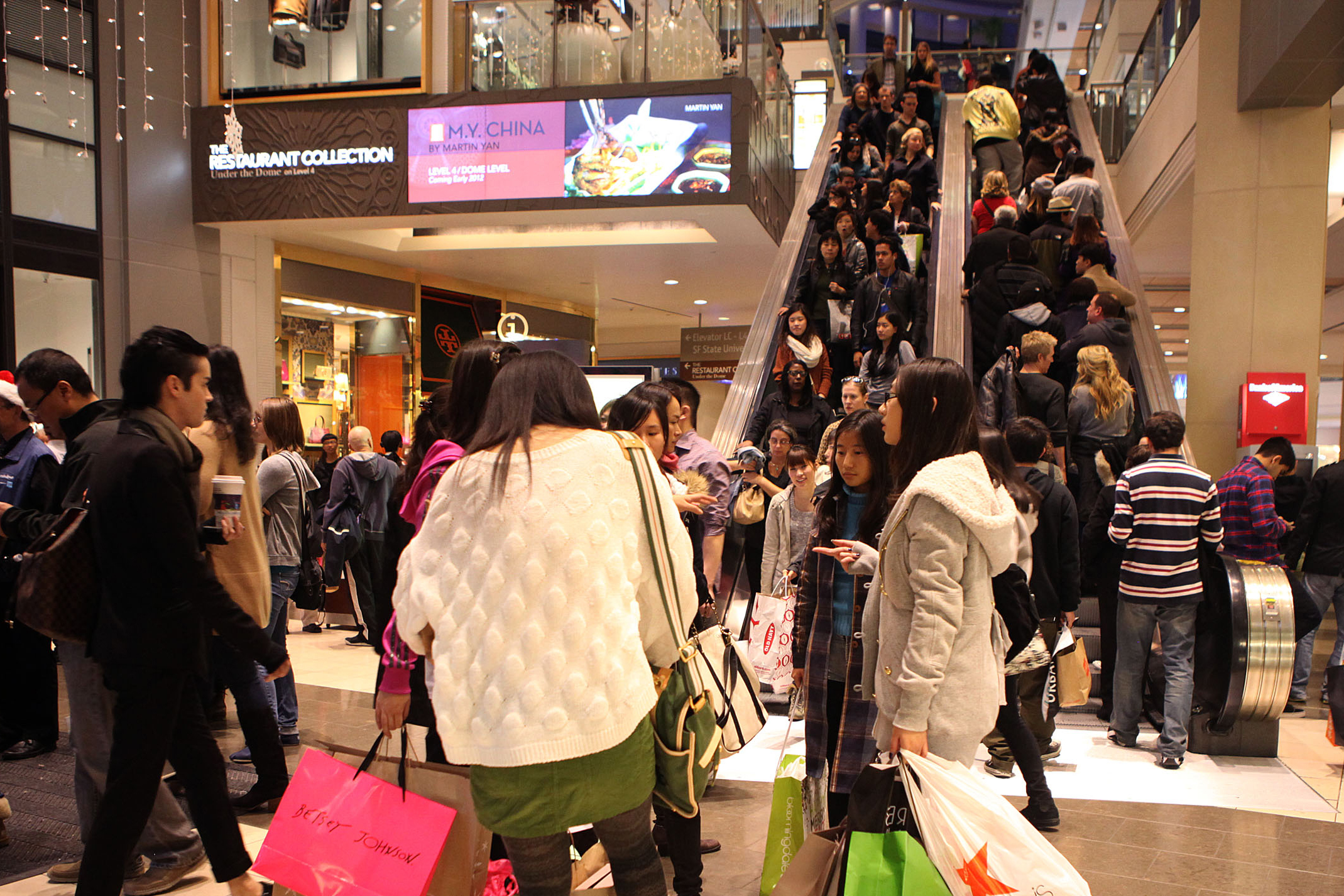 Crowded mall interior with busy escalators and many shoppers carrying bags.