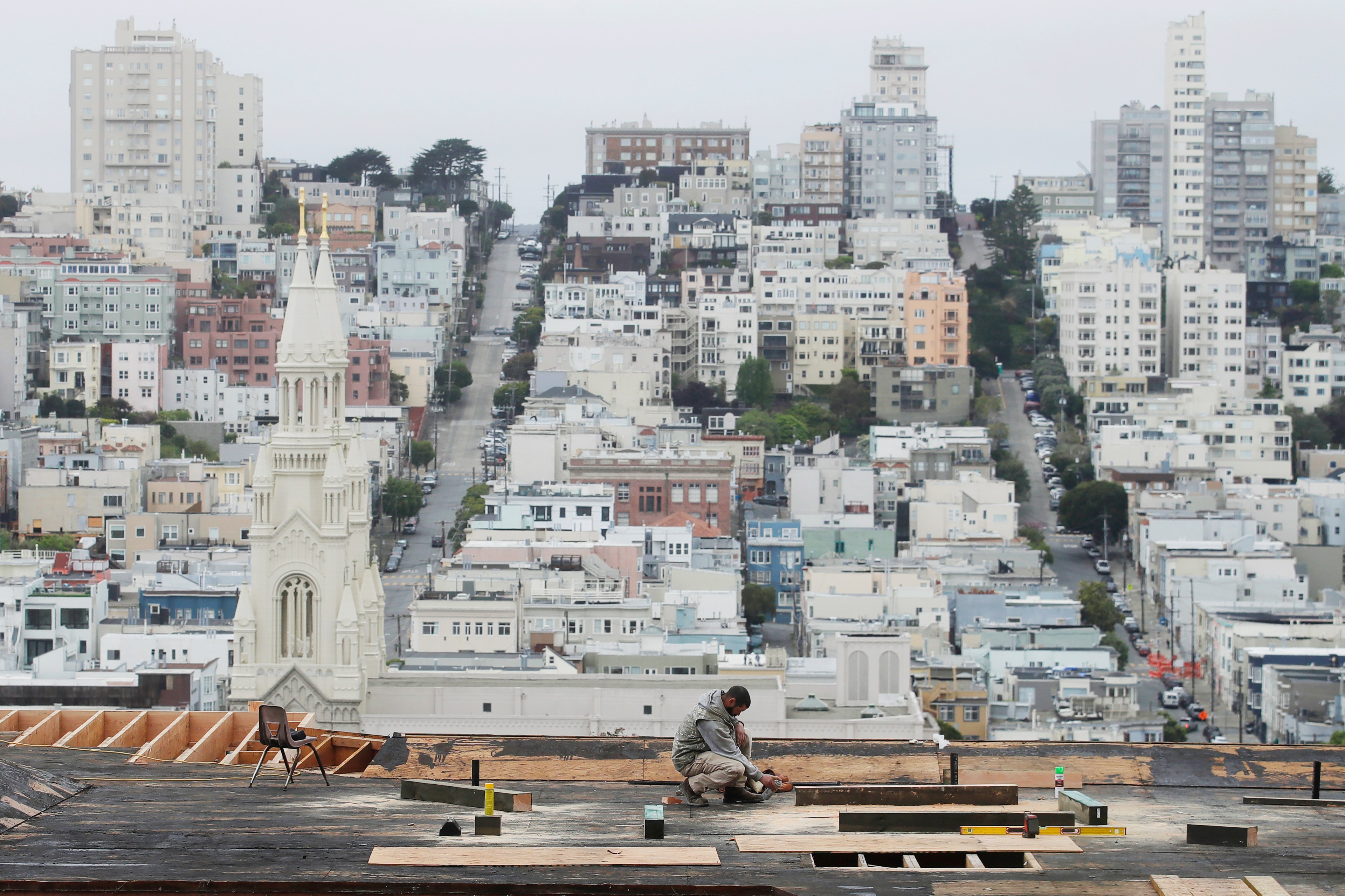 A worker on a rooftop is focused on a task, with a sprawling cityscape featuring many buildings and a prominent church spire in the background.