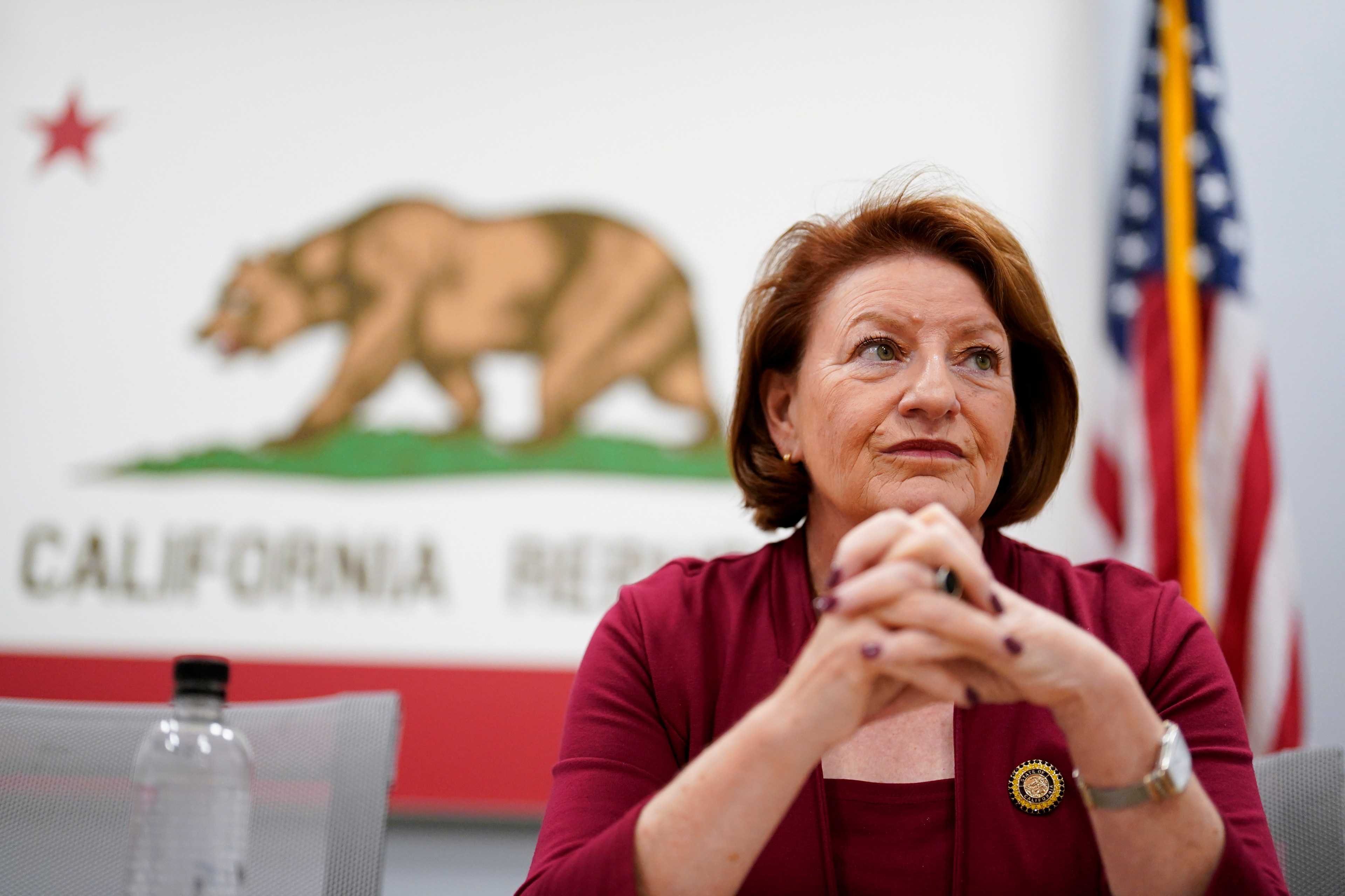 WomanWoman in red suit sits with hands clasped with a california state flag in the background