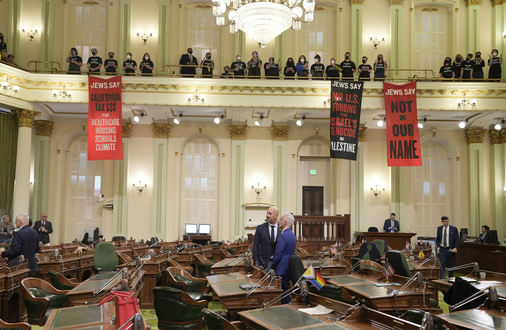 People hold protest banners in a legislative chamber with many empty seats.
