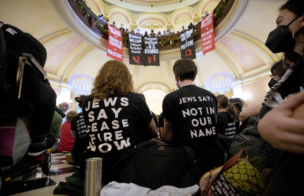 Individuals with protest shirts are inside a building with banners hanging above. The shirts read &quot;JEWS SAY CEASEFIRE NOW&quot; and &quot;NOT IN OUR NAME.&quot;