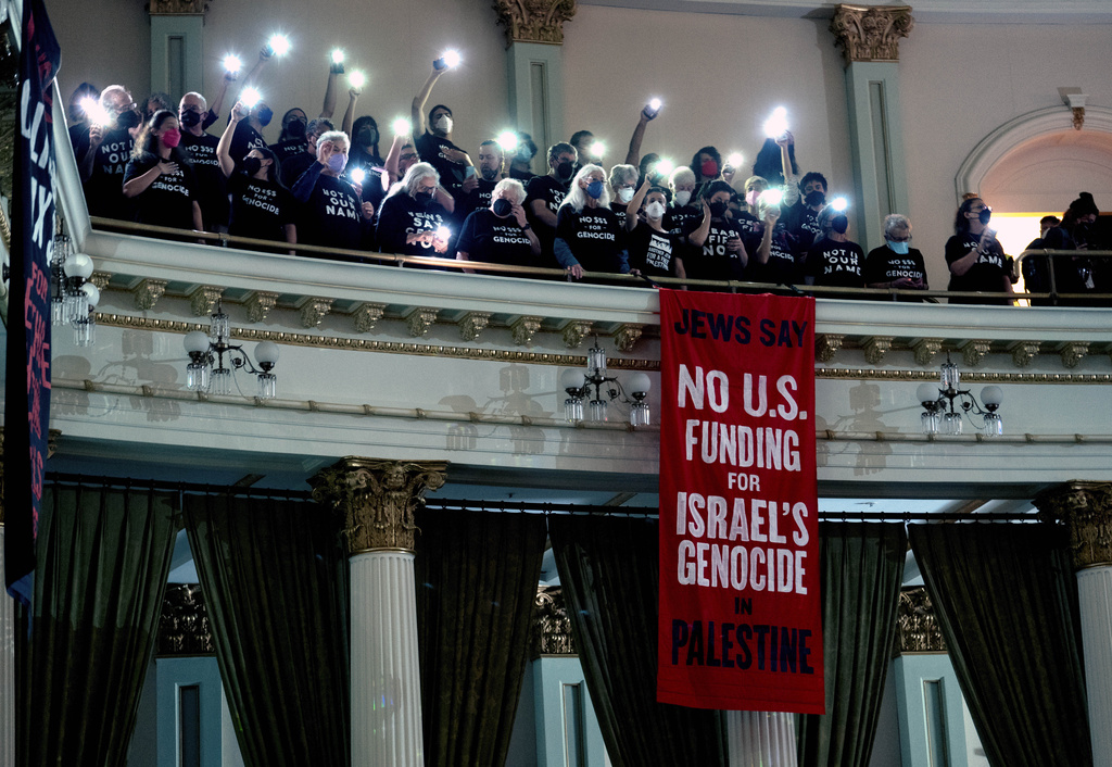 Activists with signs and lights on a balcony behind a banner protesting U.S. funding for Israel.