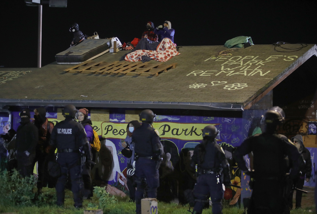 Riot police face a graffiti-covered building with protesters on its roof under a night sky. Text on the roof reads &quot;People's Park WILL REMAIN.&quot;