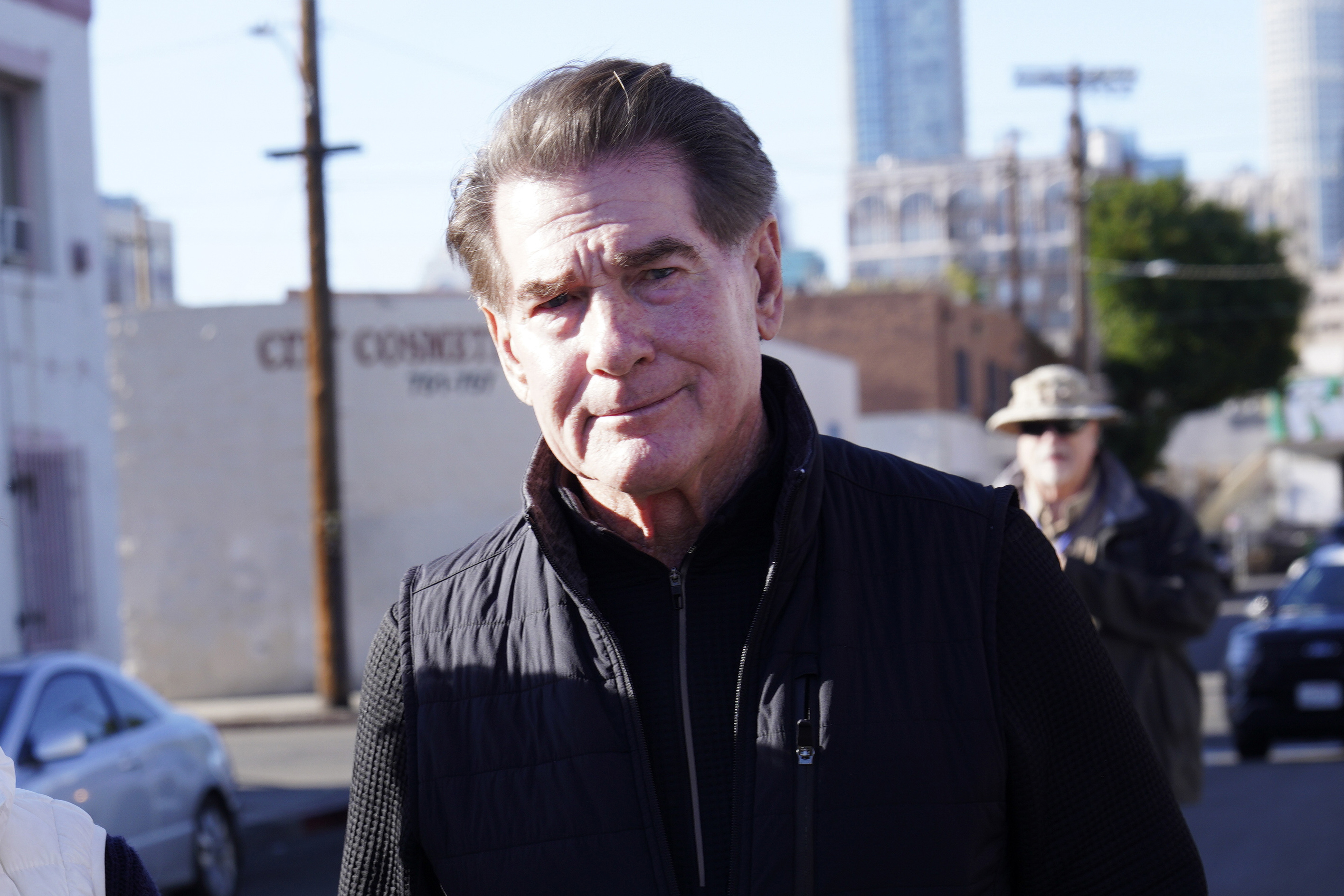 An older man with combed-back hair, wearing a black jacket, is in focus with an urban background and another person behind him.