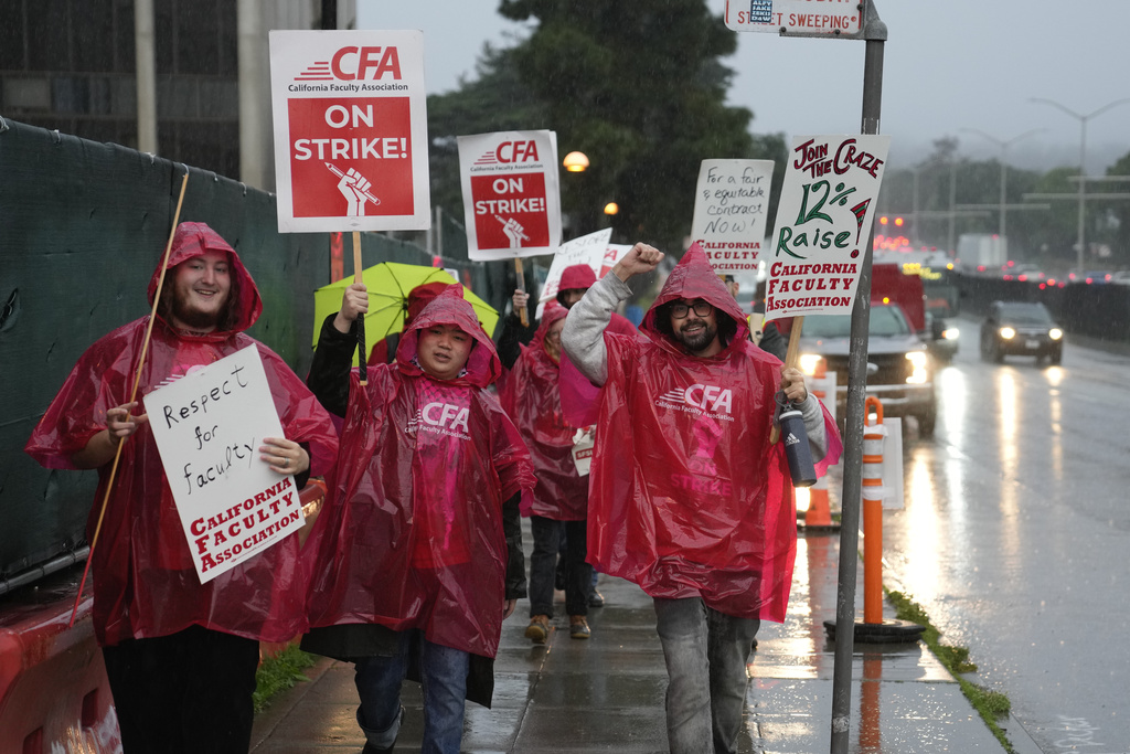 People in red rain ponchos hold strike signs by a wet road with passing cars.