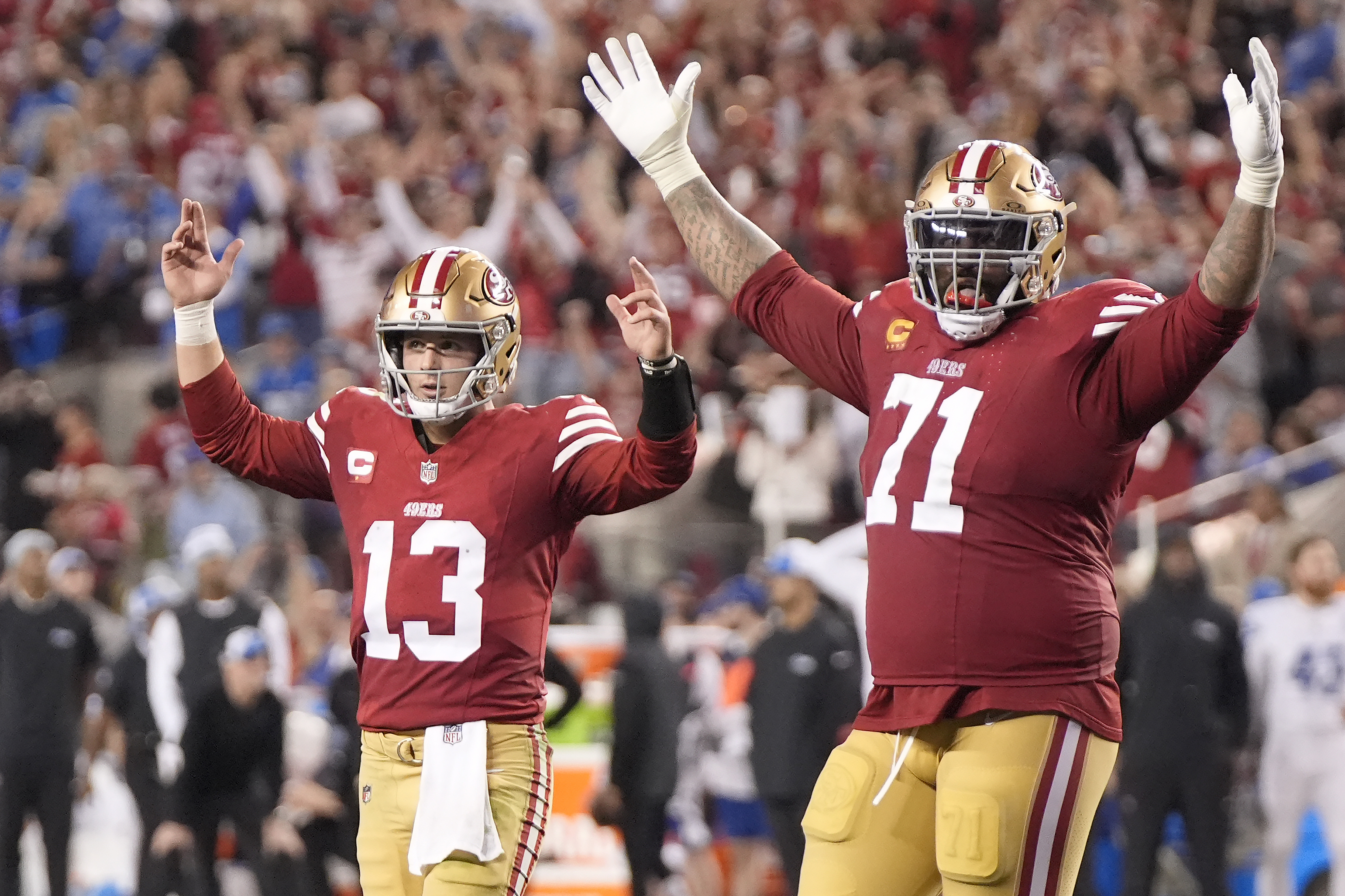 Two football players in red uniforms with '49ers' logo celebrate, raising their arms with a crowd behind them.