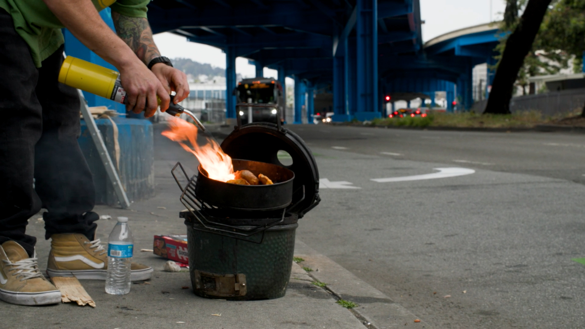 A man cooks food outdoors on a small grill using a gas torch along a sidewalk next to a street under a freeway overpass.
