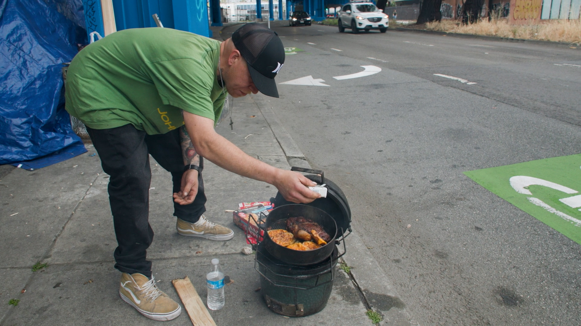 A man sprinkles salt as he cooks food outdoors on a small grill along a sidewalk next to a street under a freeway overpass.