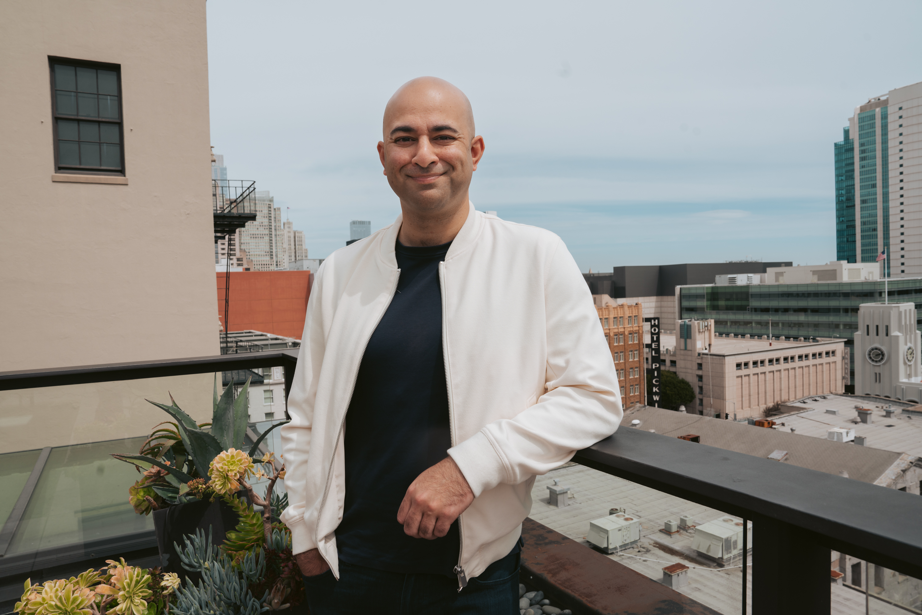 A smiling person in a white jacket stands on a balcony with urban buildings and skyscrapers in the background. There's a planter with succulents beside them.