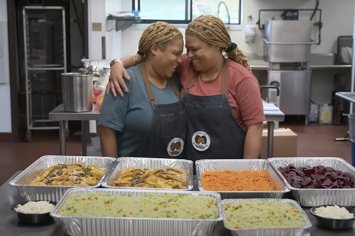 Two women stand in a kitchen hugging with pans of food in front of them.