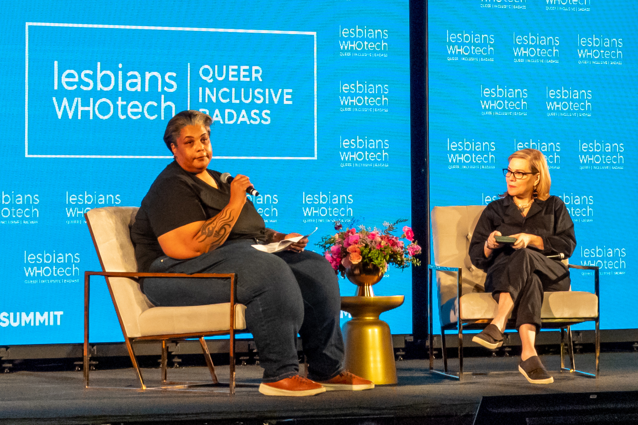 Two people sit onstage at a "Lesbians Who Tech" summit, engaged in a discussion surrounded by branding banners.
