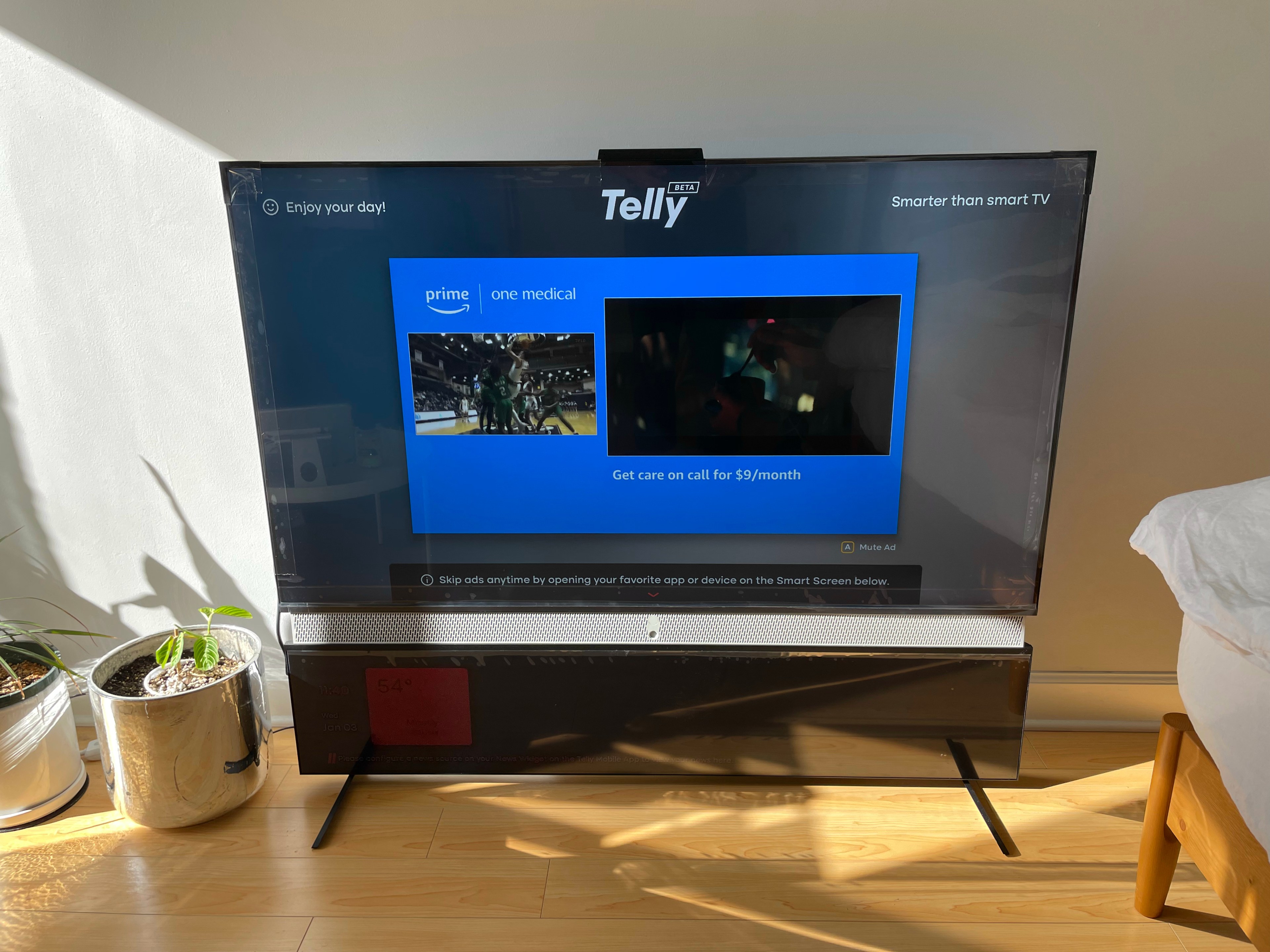 A TV displays an ad, sunlight casting shadows in the room with a plant to the left and the edge of a chair to the right.