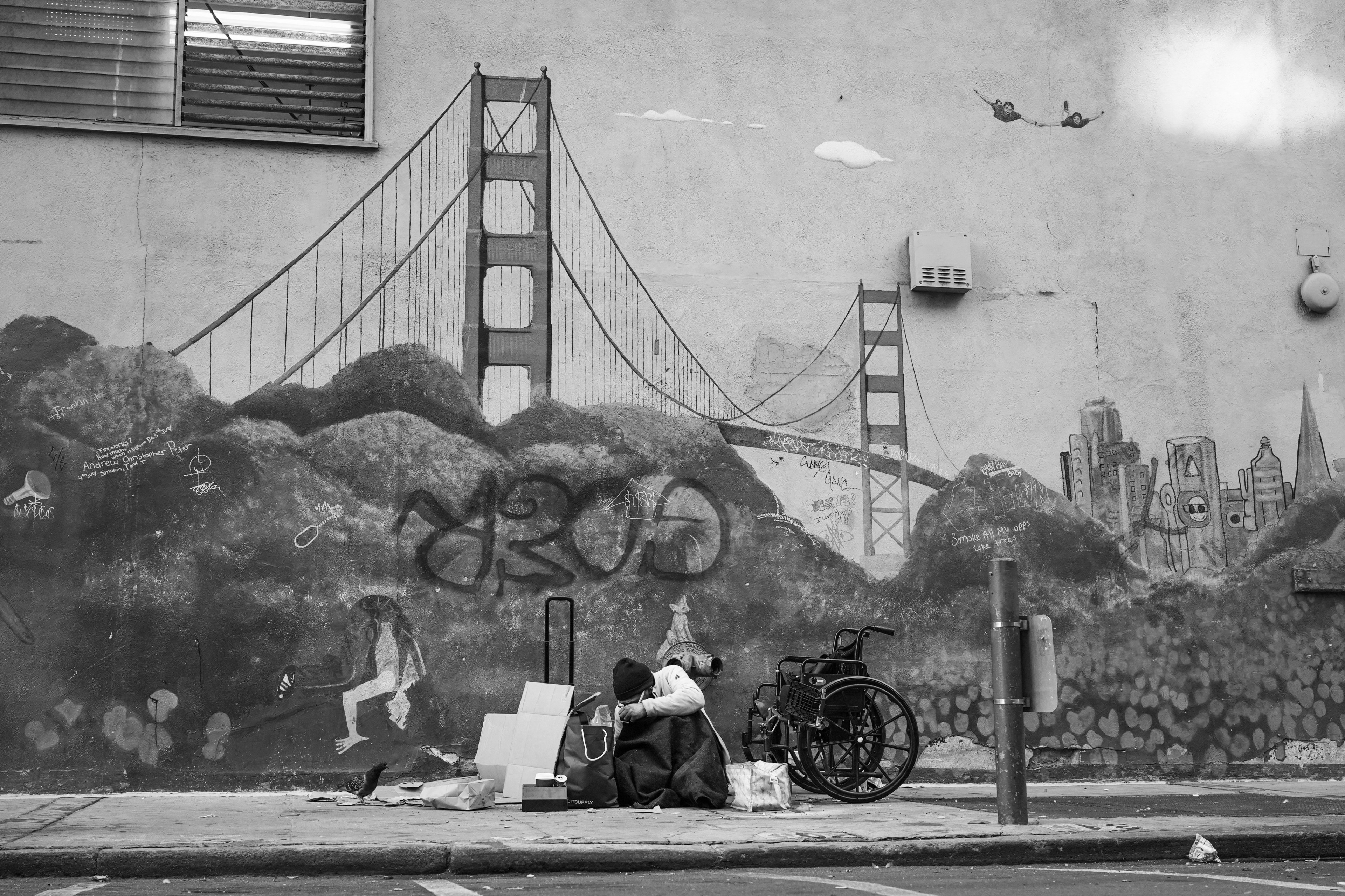 A black and white photo shows a person sitting by cardboard boxes on a sidewalk, with a mural of the Golden Gate Bridge and cityscape behind them.