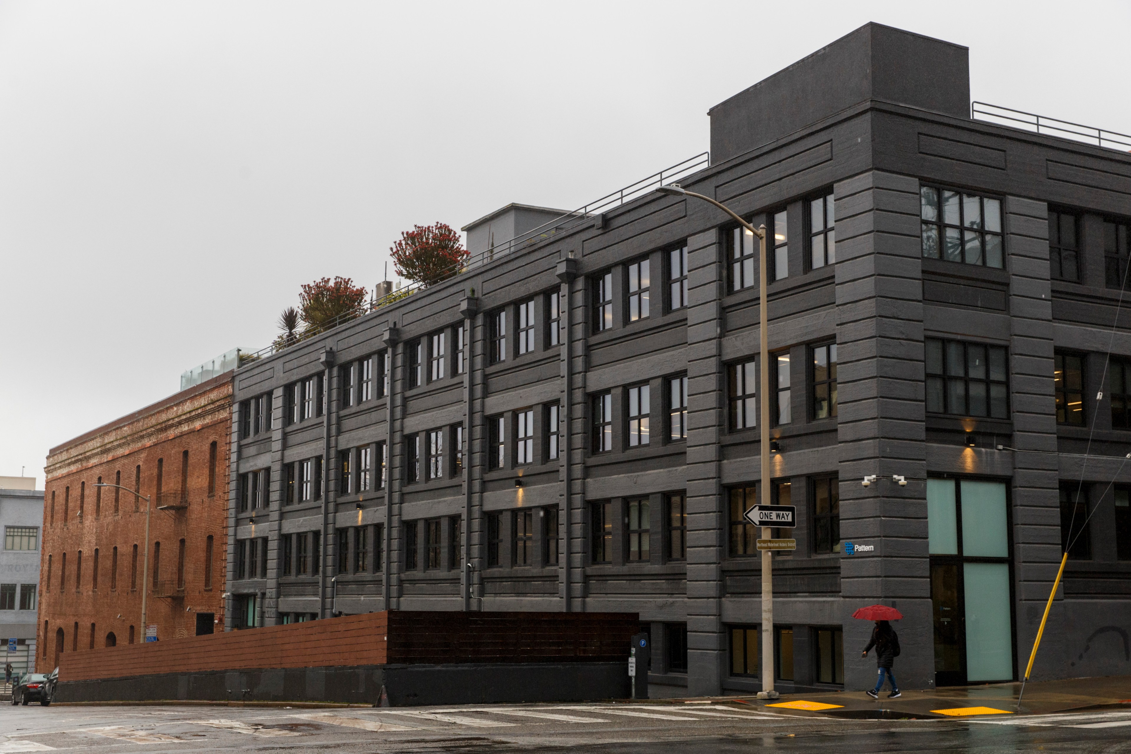 A modern gray building next to an older brick one on a wet street, with a person holding a red umbrella walking by.