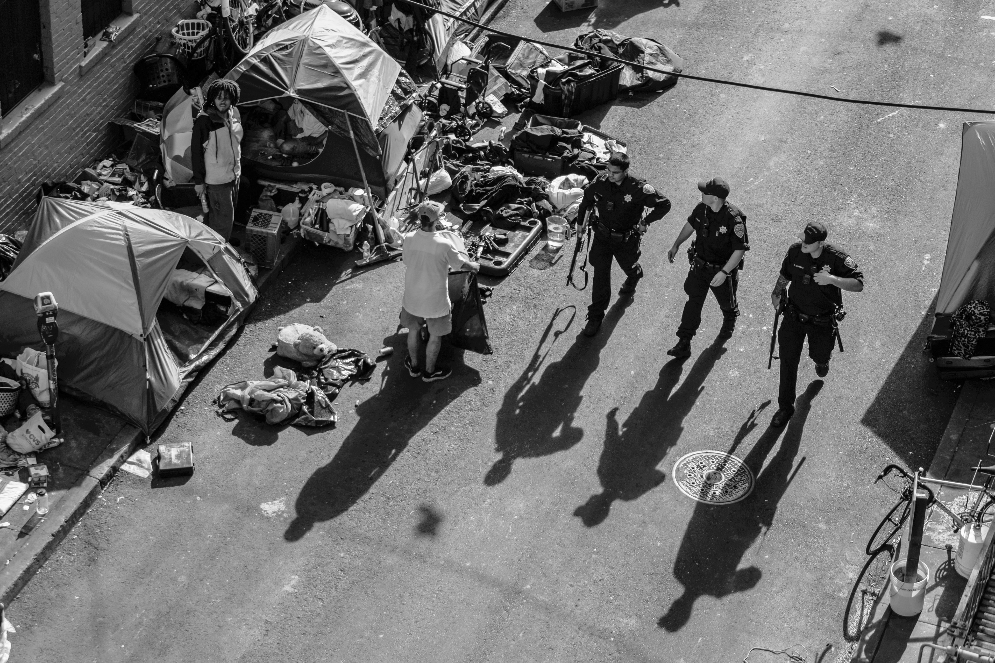 An overhead shot of a San Francisco street with tents, scattered items, and three officers standing together casting long shadows.