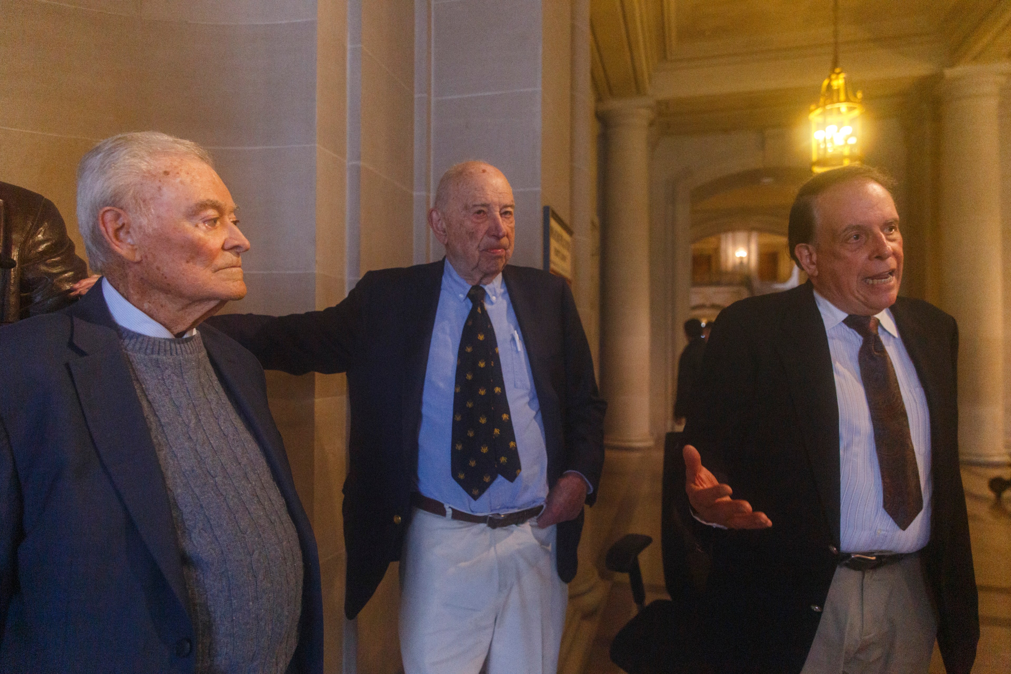 Three elderly men in suits are gathered in a room with classical architecture and warm lighting.