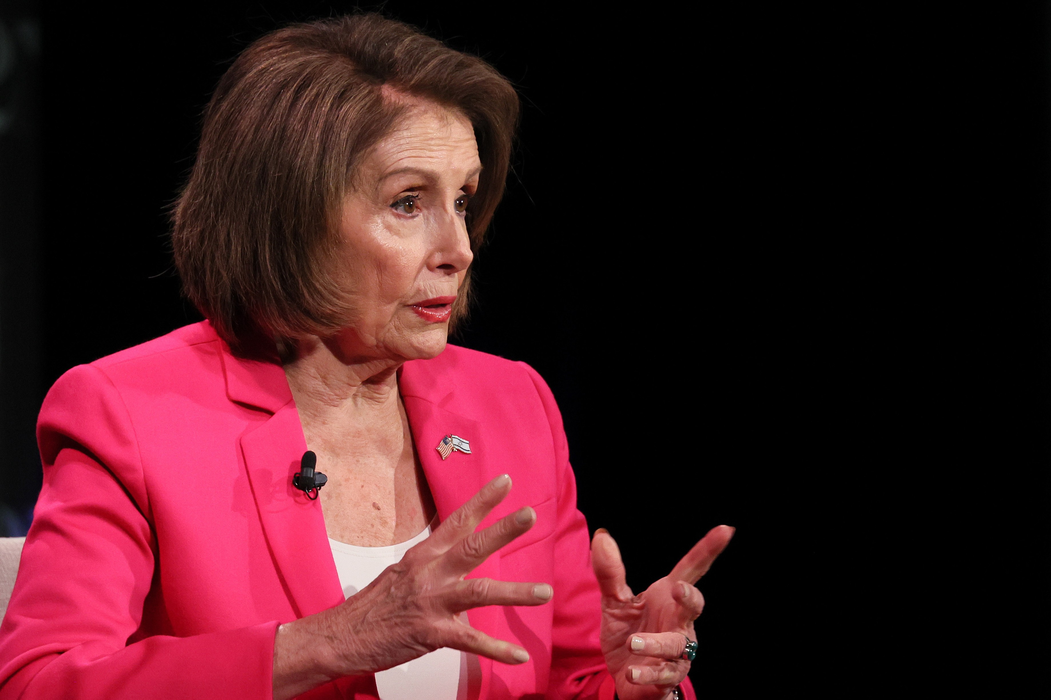 A woman in a pink blazer speaks animatedly, wearing a pin and a mic, against a dark background.