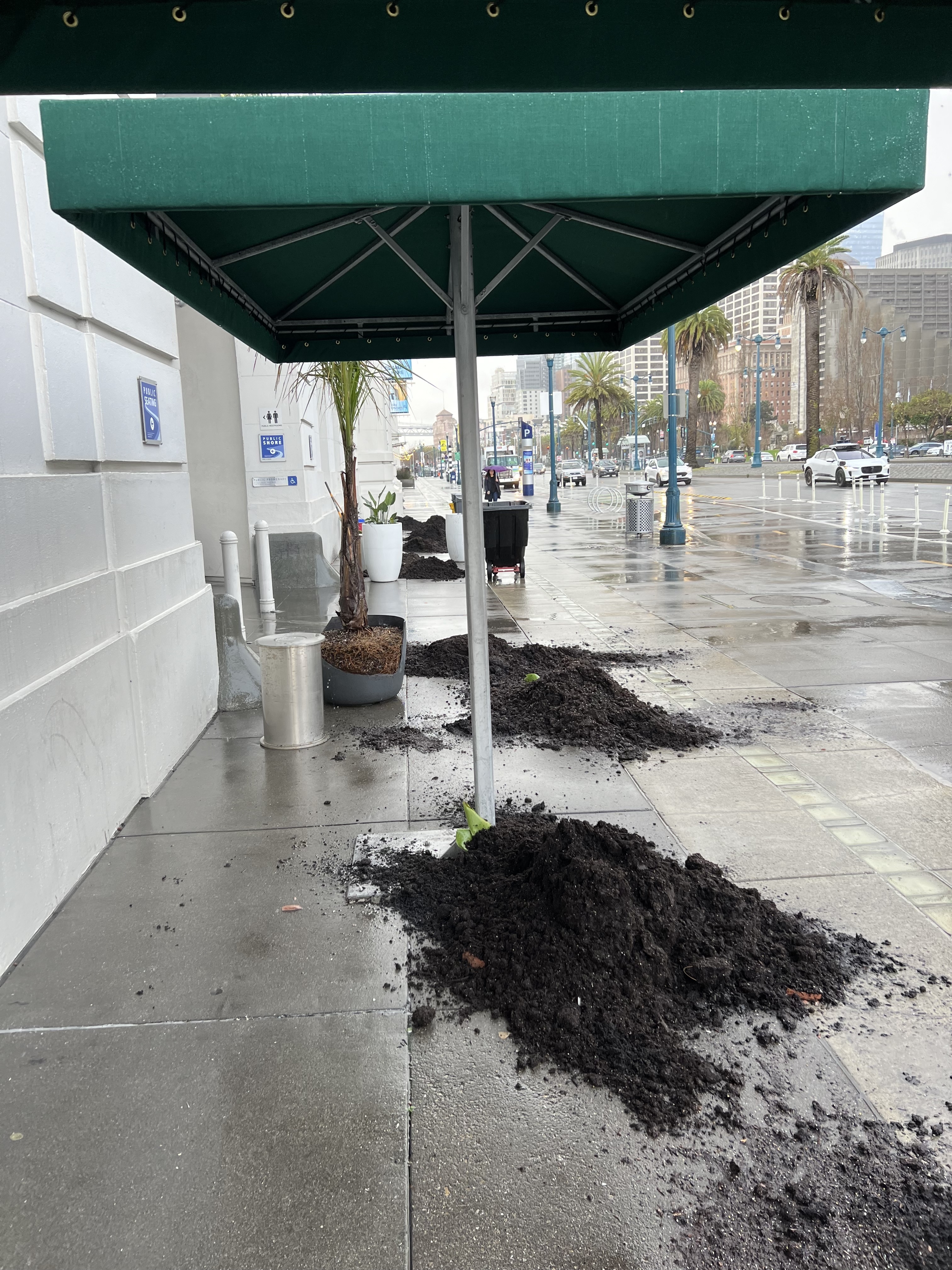 A wet sidewalk under a green awning with scattered piles of overturned planter soil.