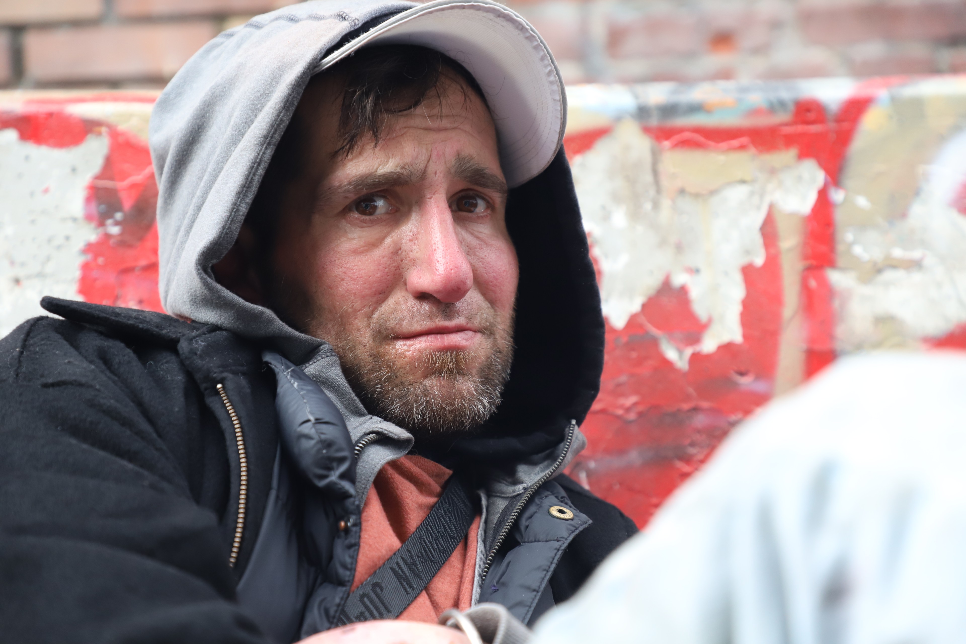 A man in layers with a hood, a beard, and a concerned expression sits before a peeling red wall.