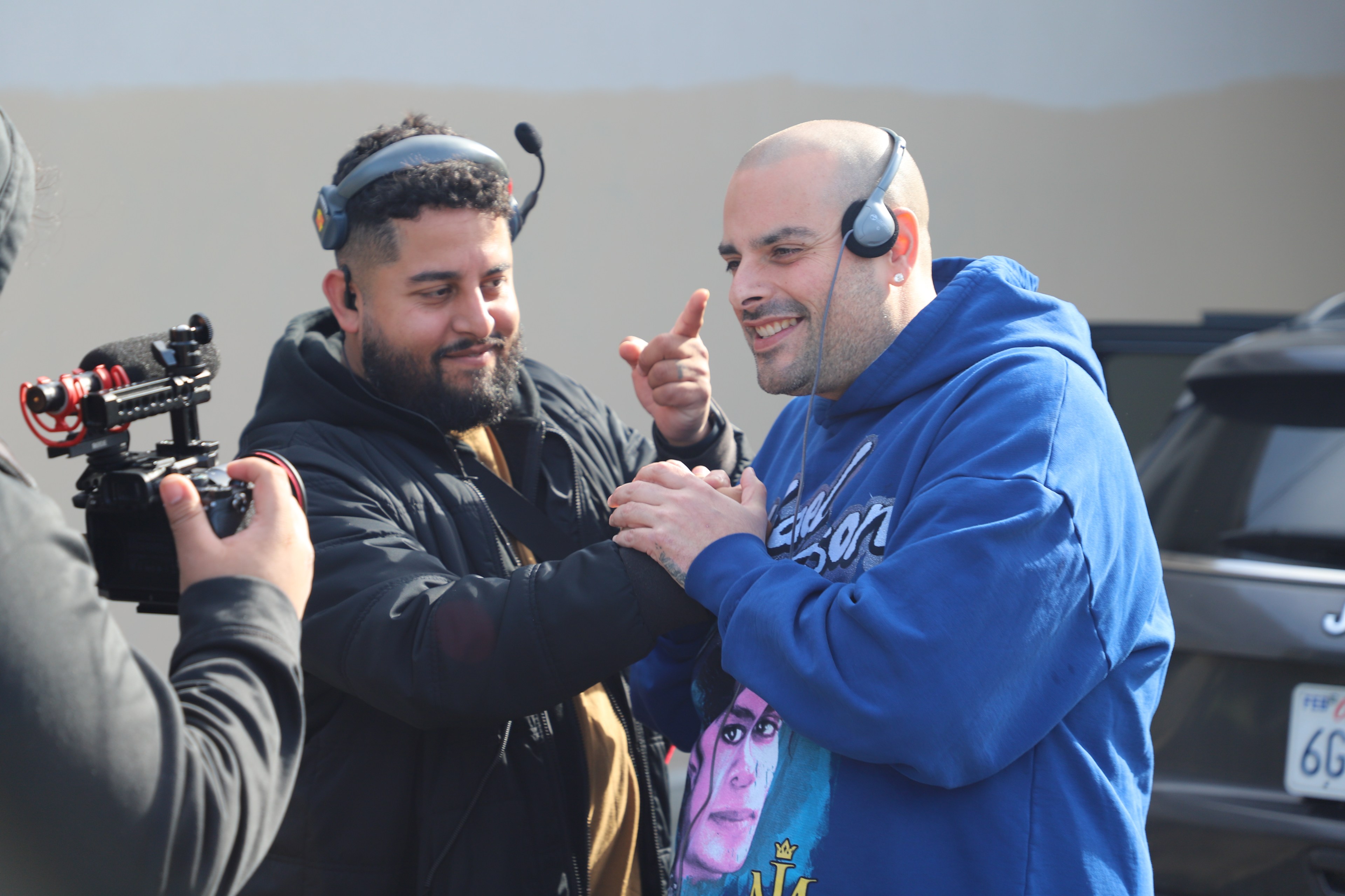 Two men are interacting joyfully outdoors; one points a camera at the other, who gestures and smiles, wearing headphones and a blue hoodie.