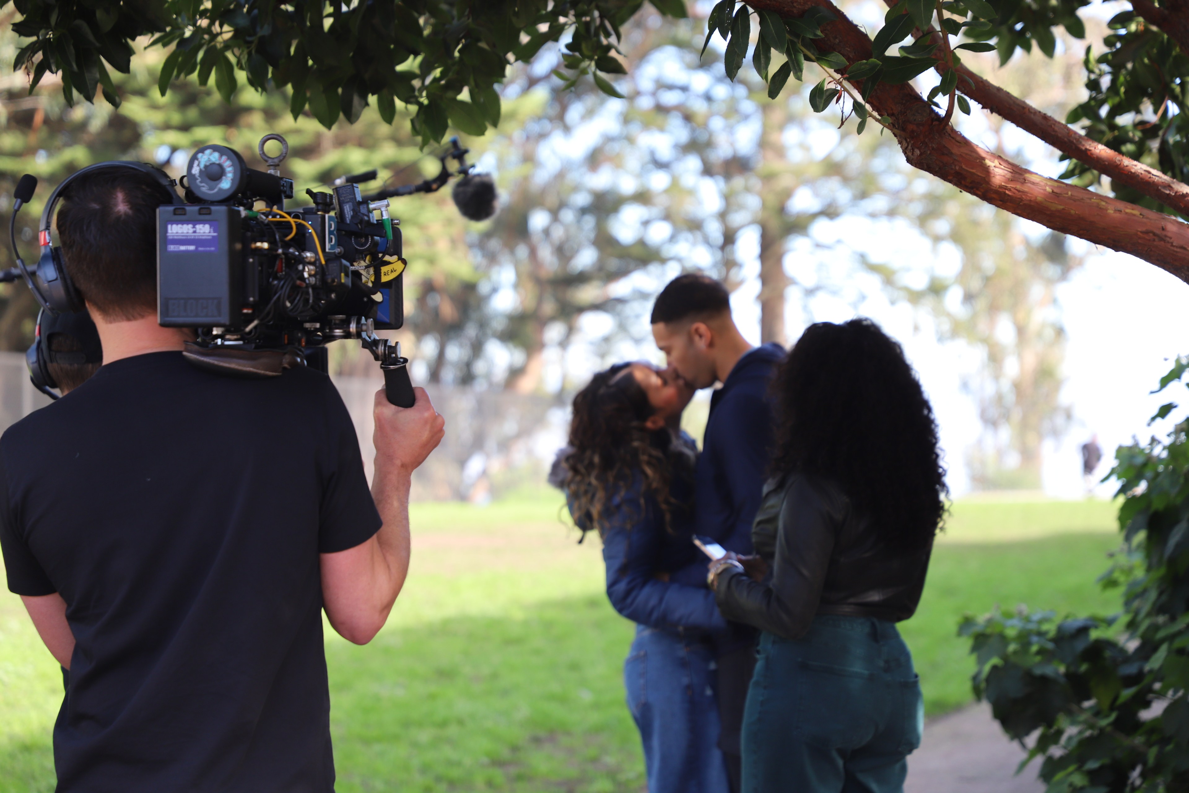A cameraman films a couple kissing in a park, while another person watches, surrounded by trees.